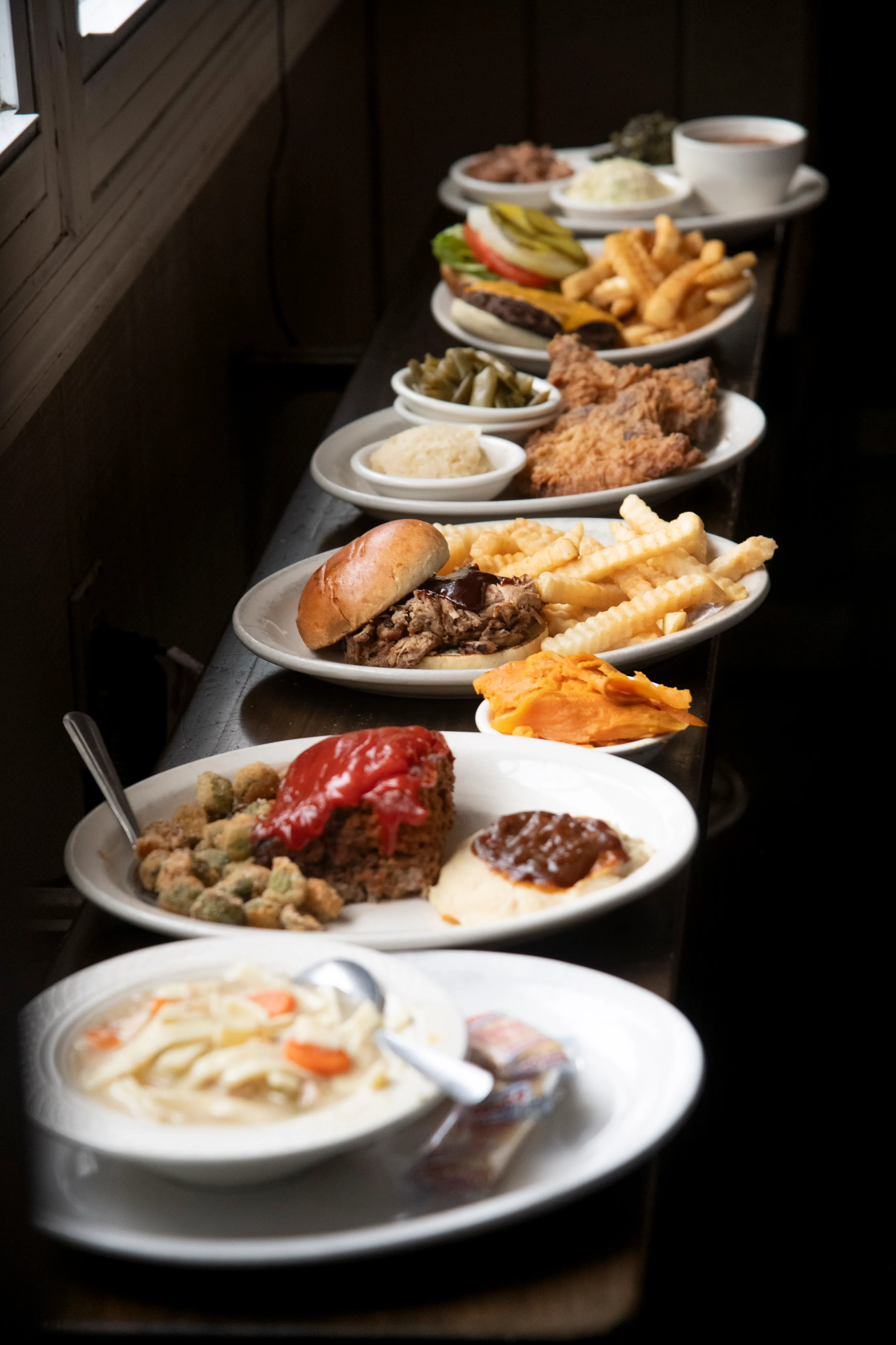 A variety of plated meals including chicken noodle soup, meatloaf with mashed potatoes and fried okra, pulled pork sandwich with fries, fried chicken with green beans and mashed potatoes, cheeseburger with fries, and sides of cheese slices, served on a wooden surface near a window.