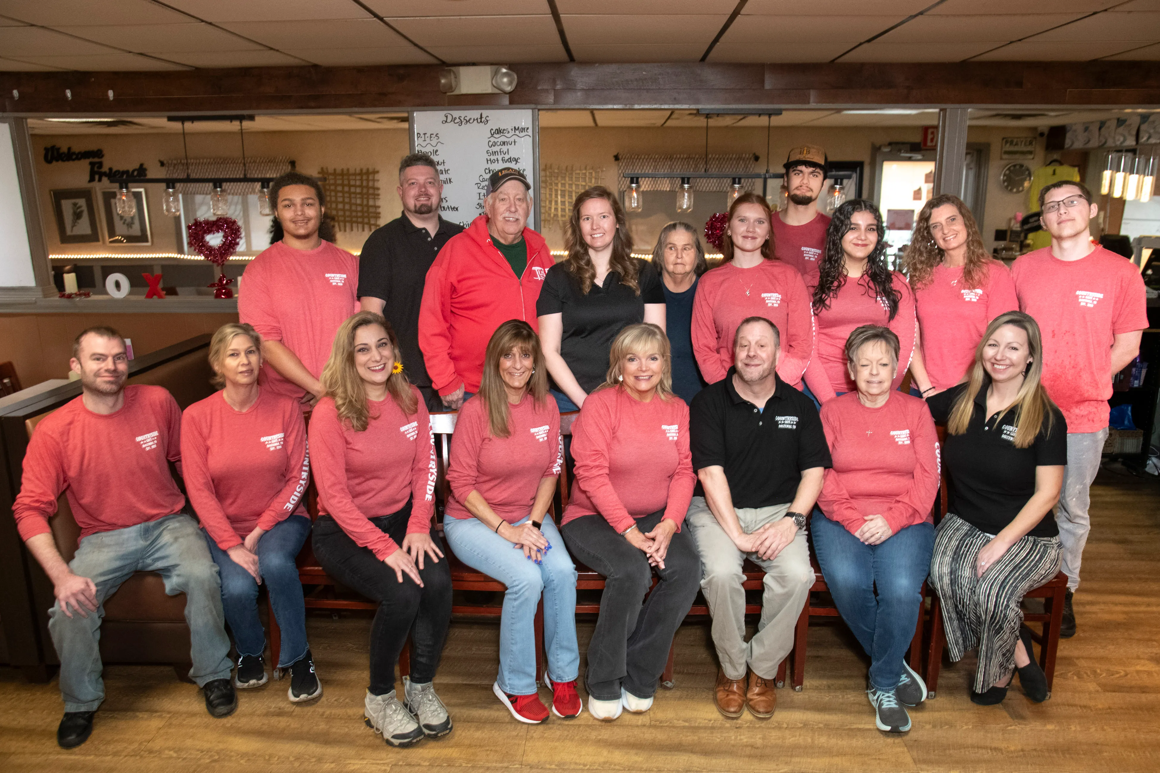 Group portrait of 20 people, mostly wearing red long-sleeve shirts with white text, seated and standing inside a restaurant with a dessert menu visible in the background.
