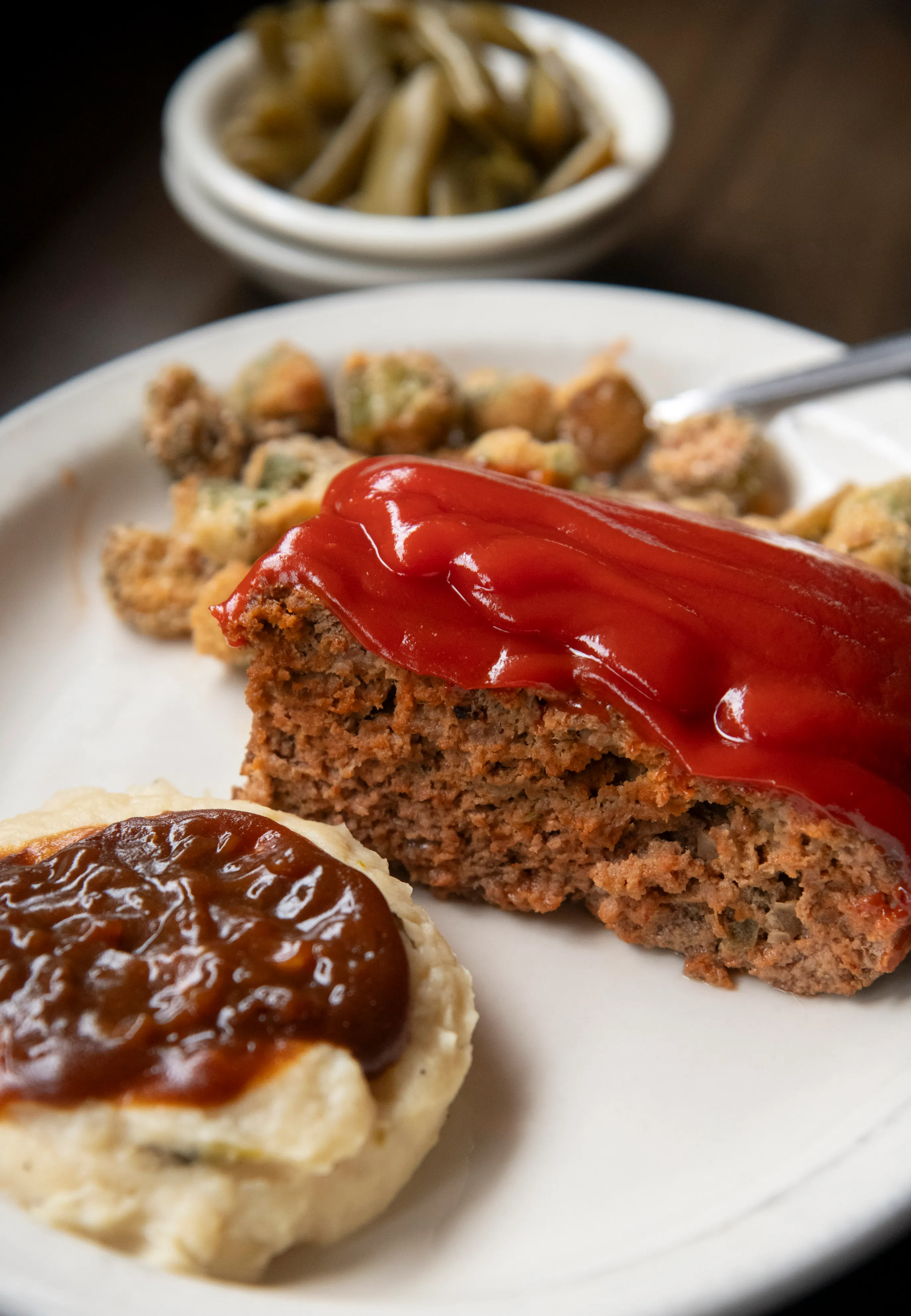 Plate with a slice of meatloaf topped with ketchup, mashed potatoes with brown gravy, and fried okra, with a bowl of green beans in the background.