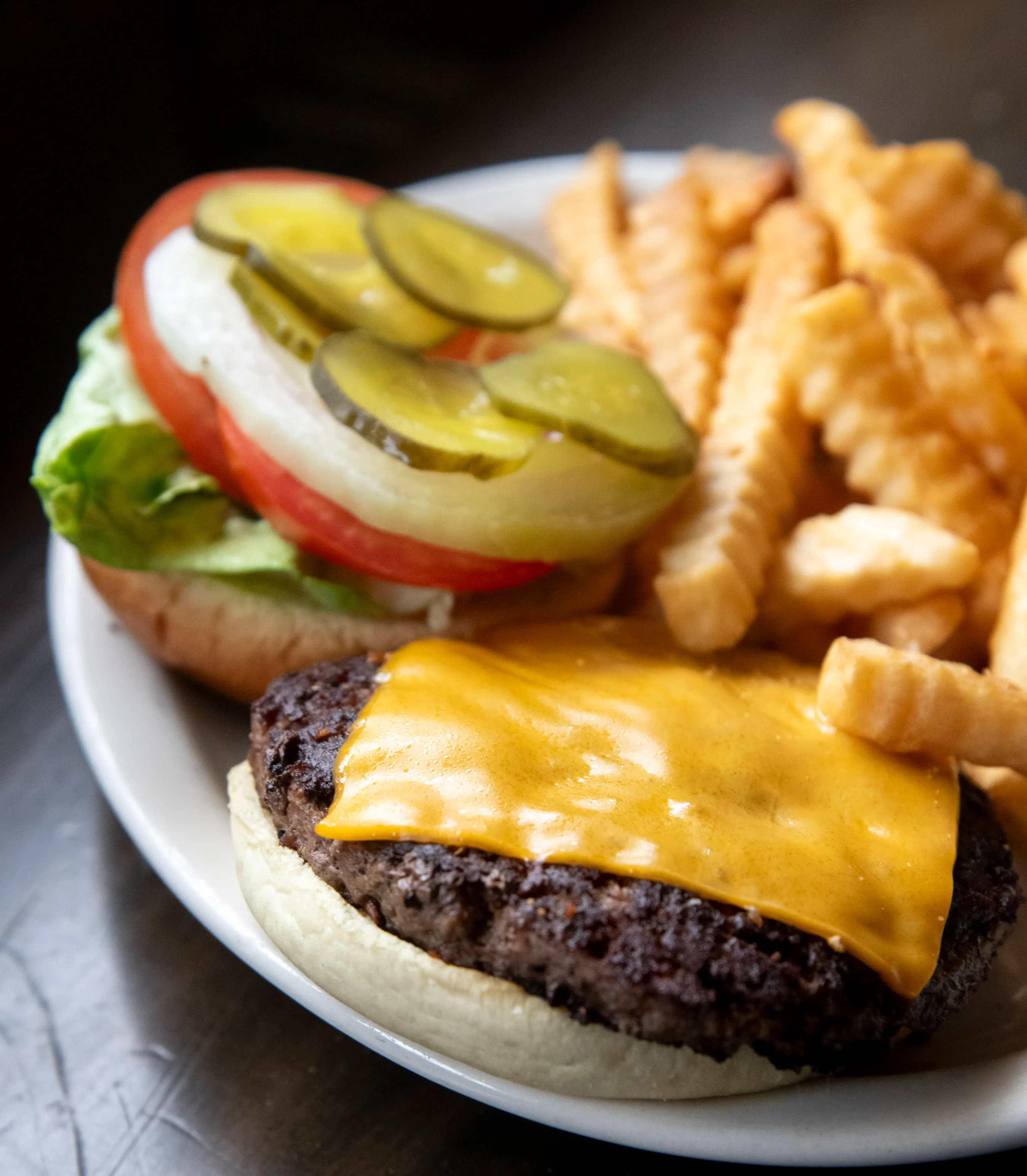 Cheeseburger with pickles, onions, tomato, and lettuce on a bun served with crinkle-cut fries on a white plate.