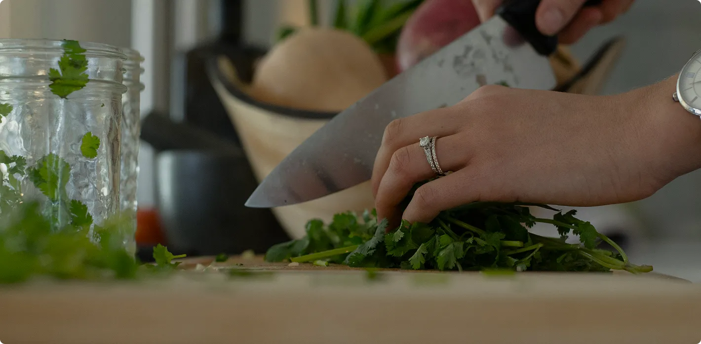 Close-up of hands chopping fresh cilantro on a wooden cutting board with a large kitchen knife.