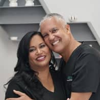 Smiling man and woman in black outfits embracing indoors under gray ceiling beams.