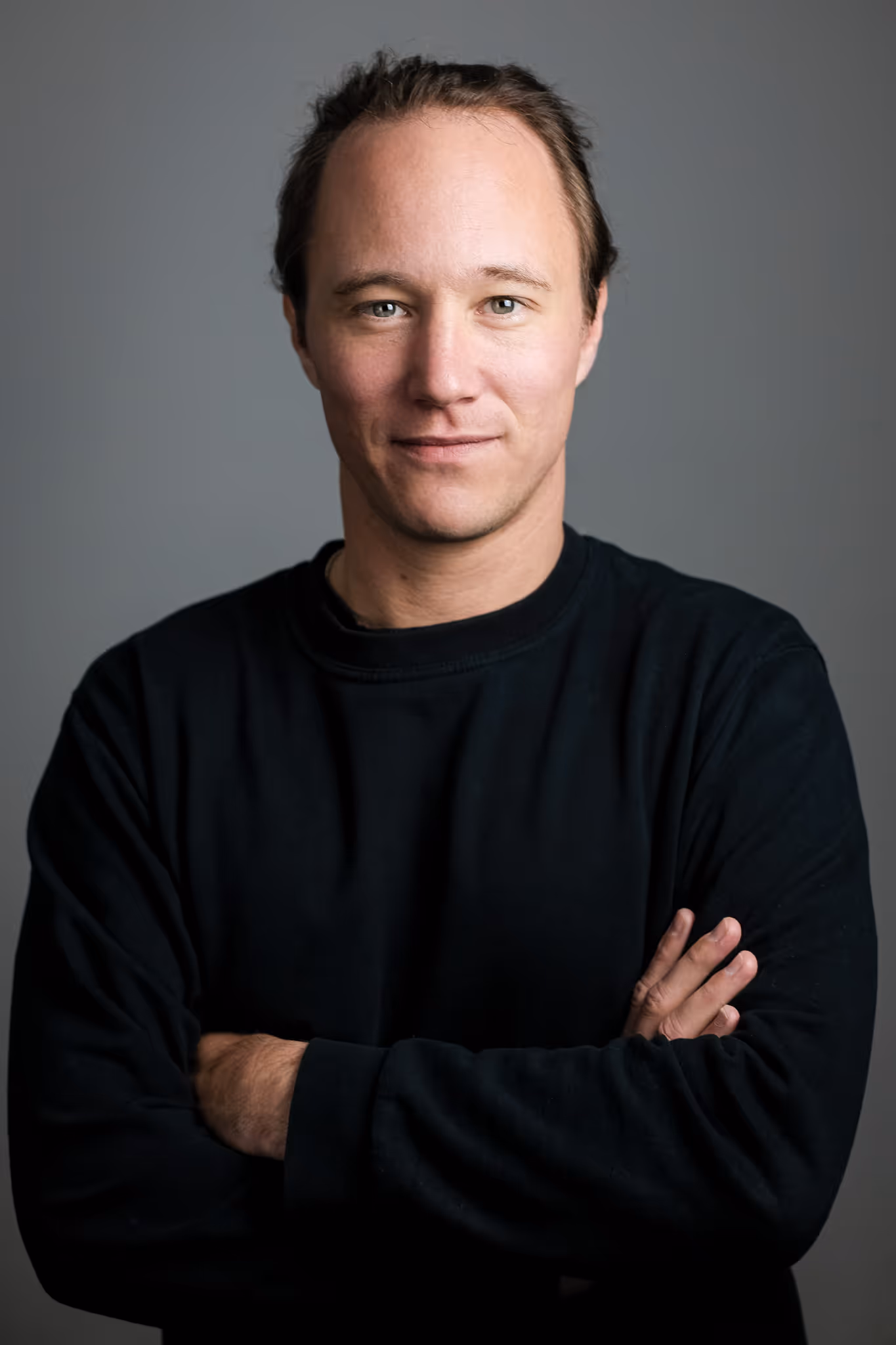 Max with light skin and light brown hair tied back, wearing a black long-sleeve shirt, standing with arms crossed against a plain gray background.