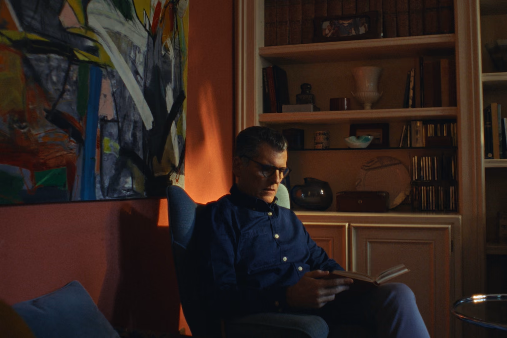 Man in glasses sitting in armchair reading a book in a dim room with abstract art and shelves.