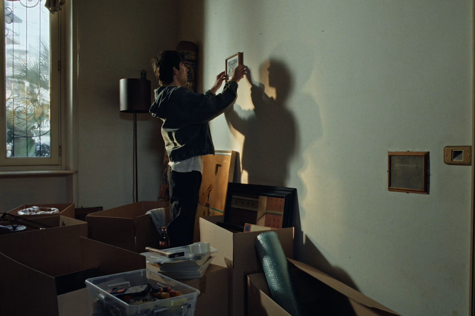 Person hanging a framed picture on a wall surrounded by unpacked cardboard boxes and packing materials.