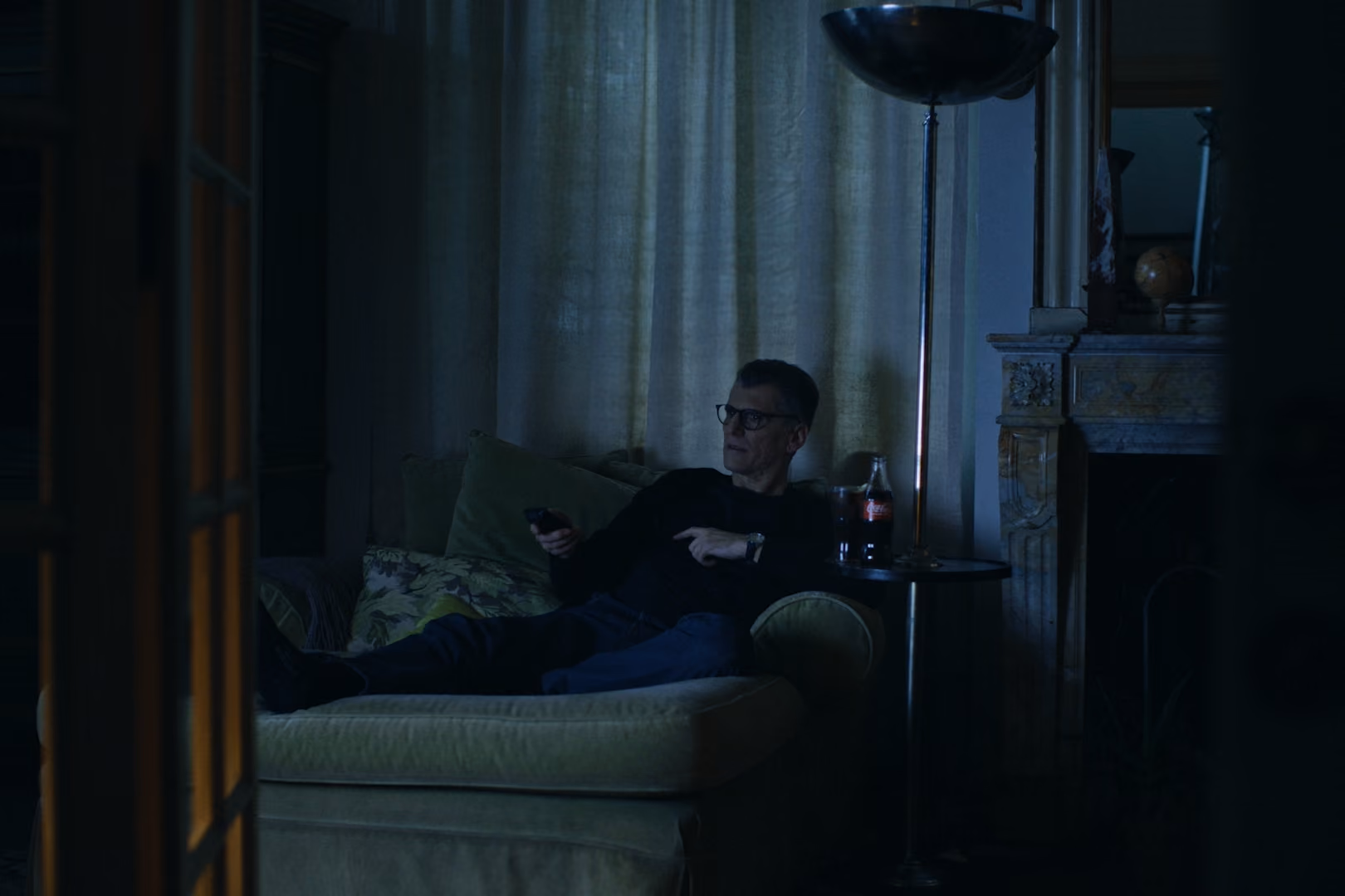 Man wearing glasses lounging on couch holding remote with bottle of Coke on side table.