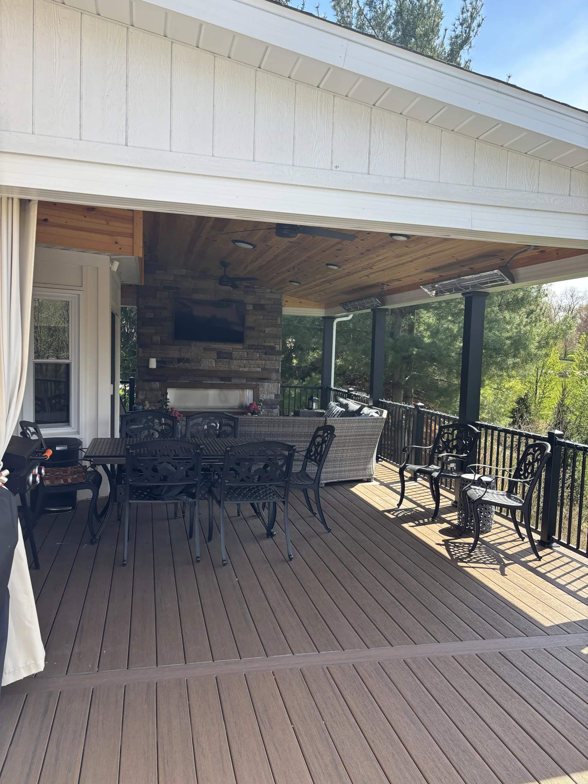 covered back deck with black table and chairs. And a couch facing a wall-mounted TV on a stone wall