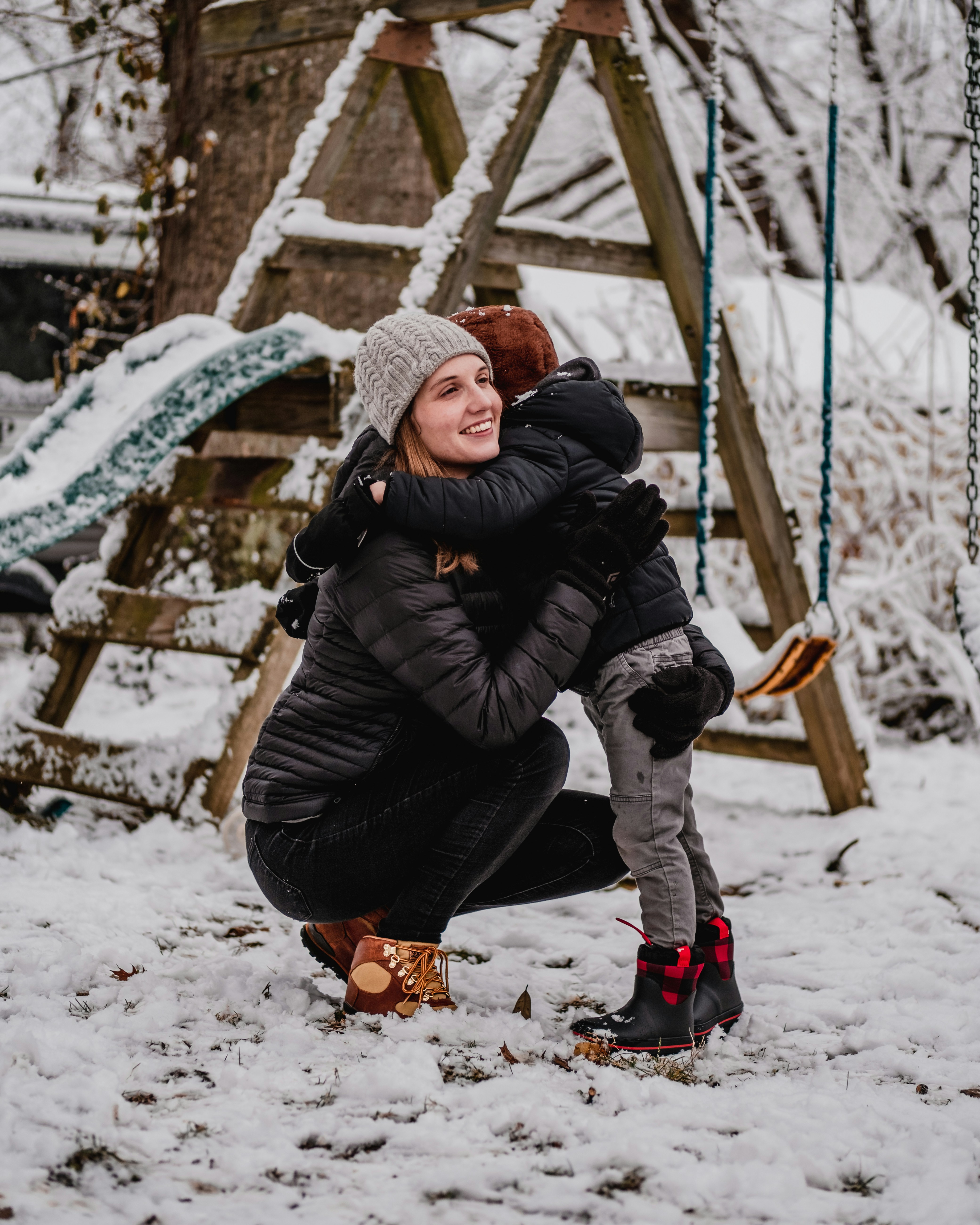 Nanny holding a child outdoors near a snow-covered play structure during winter weather