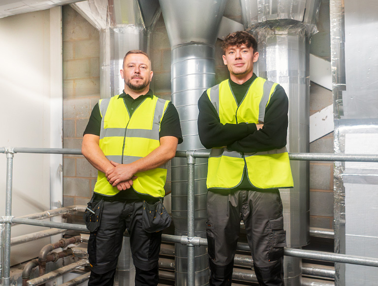 Two men in high-viz jackets standing in front of metal pipes