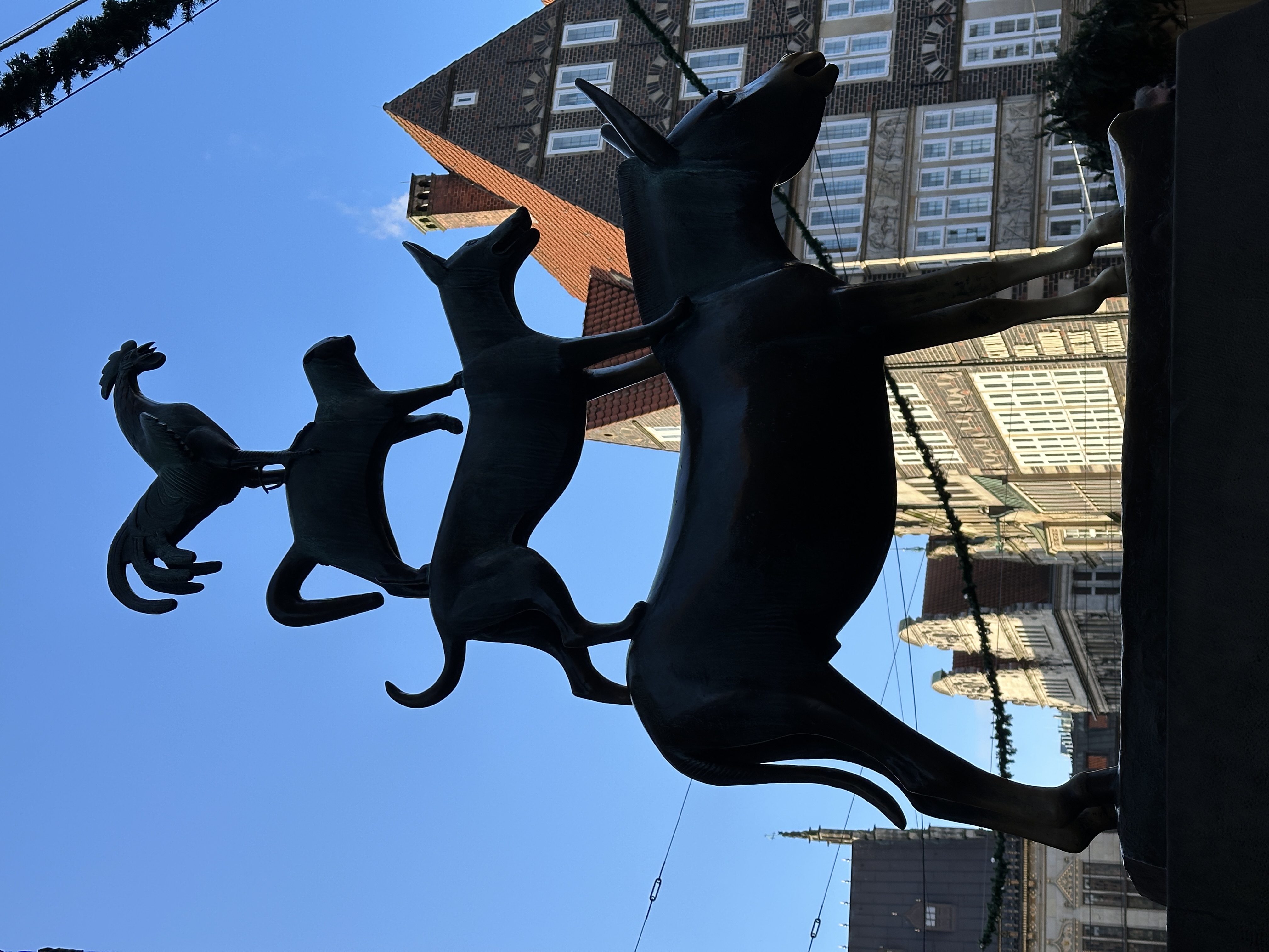 Silhouetted statue of the Bremen Town Musicians—a rooster standing on a cat, which is standing on a dog, which is standing on a donkey—with historic buildings in the background under a clear blue sky.