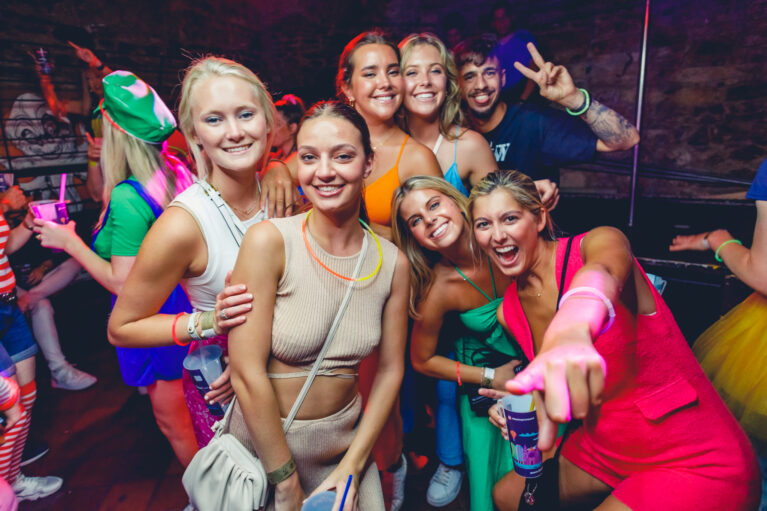 Group of seven young adults smiling and posing together at a lively indoor party with colorful outfits and neon accessories.