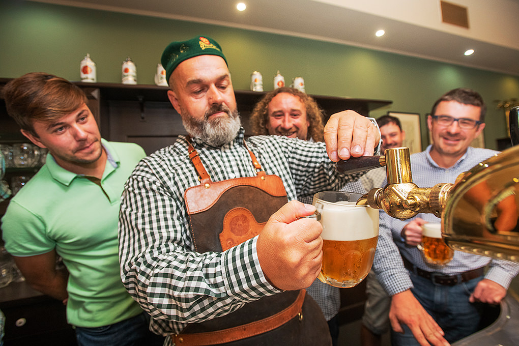 Bartender in a green checkered shirt and leather apron pouring beer from a tap while four men watch and smile.