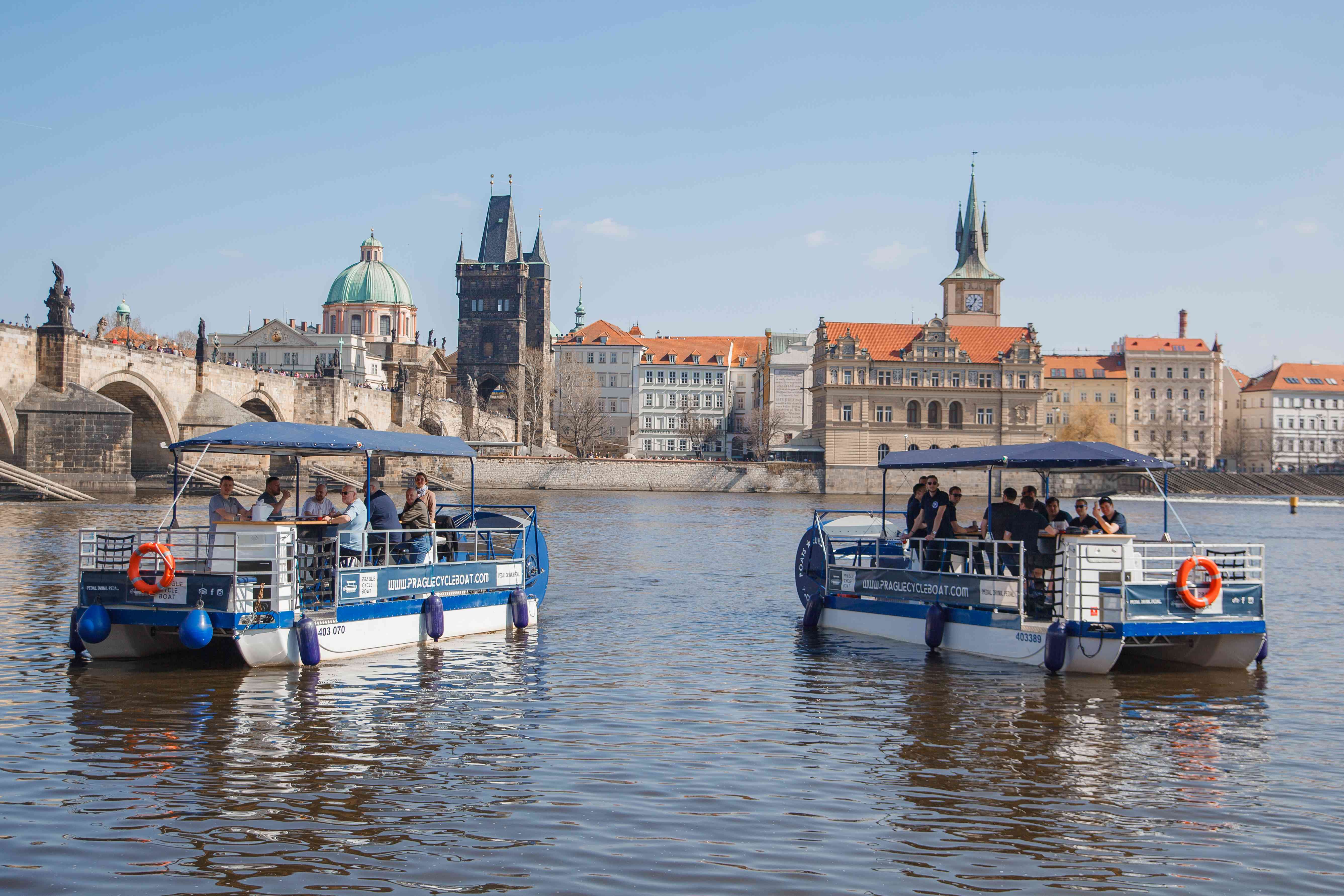 Two blue pedal boats with passengers on a river in Prague, with historic buildings and Charles Bridge in the background.