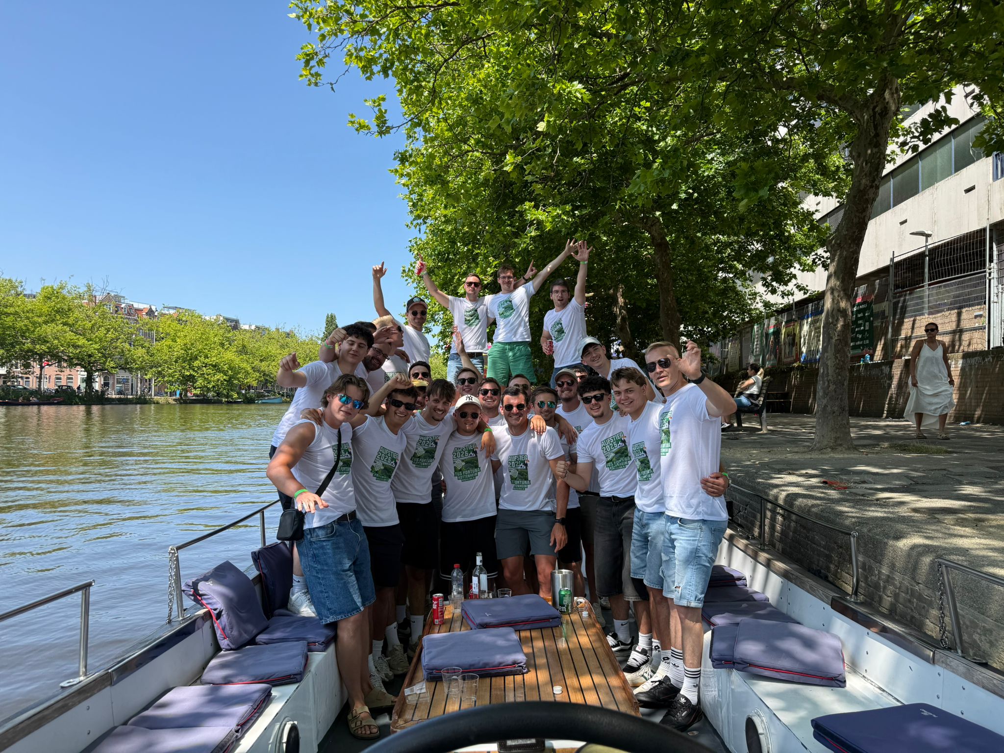 Group of young men in matching white T-shirts posing and smiling on a boat on a sunny day by a riverside with trees and buildings in the background.