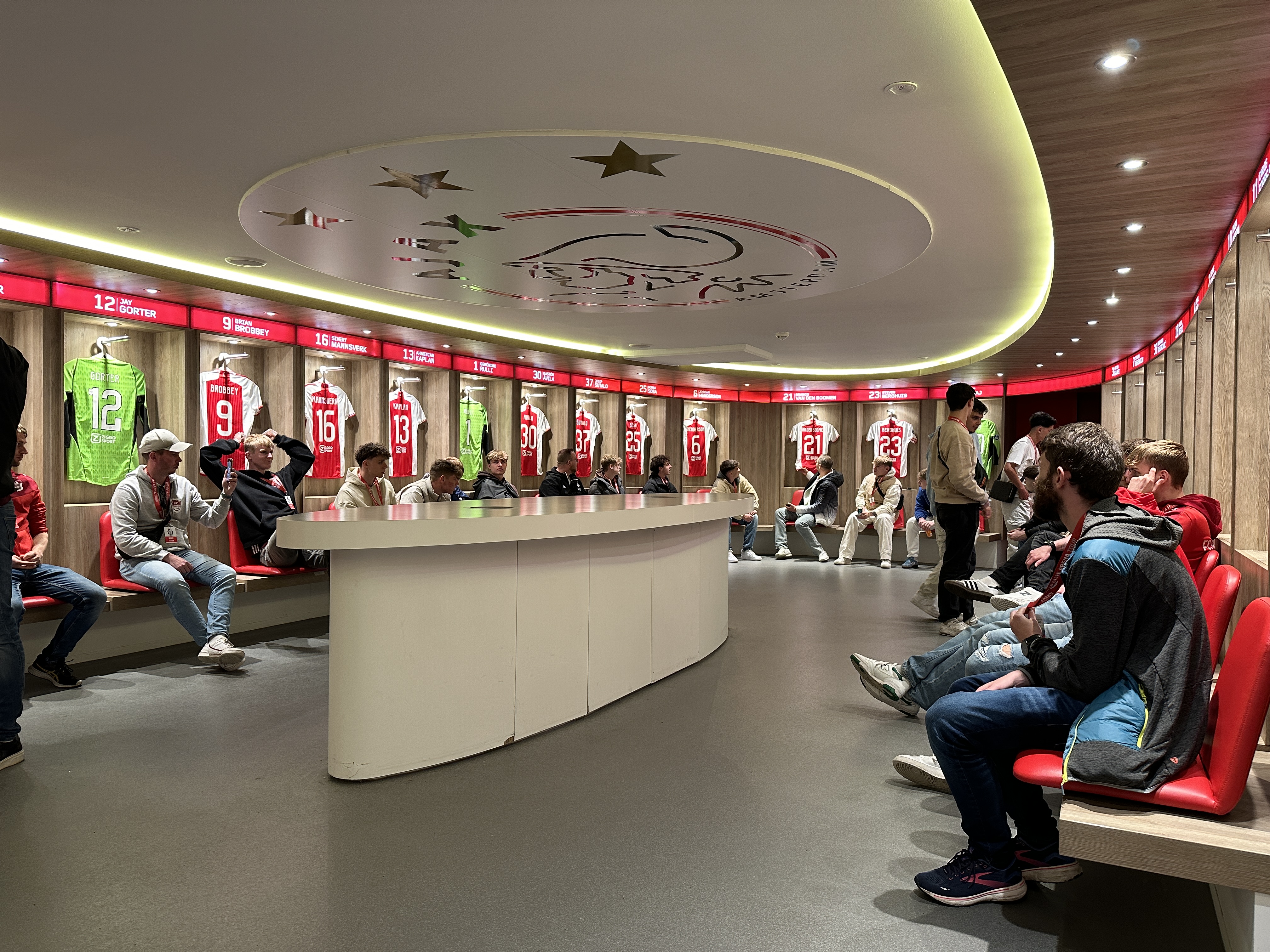 Locker room with red and white Ajax soccer jerseys displayed above seated people on benches.