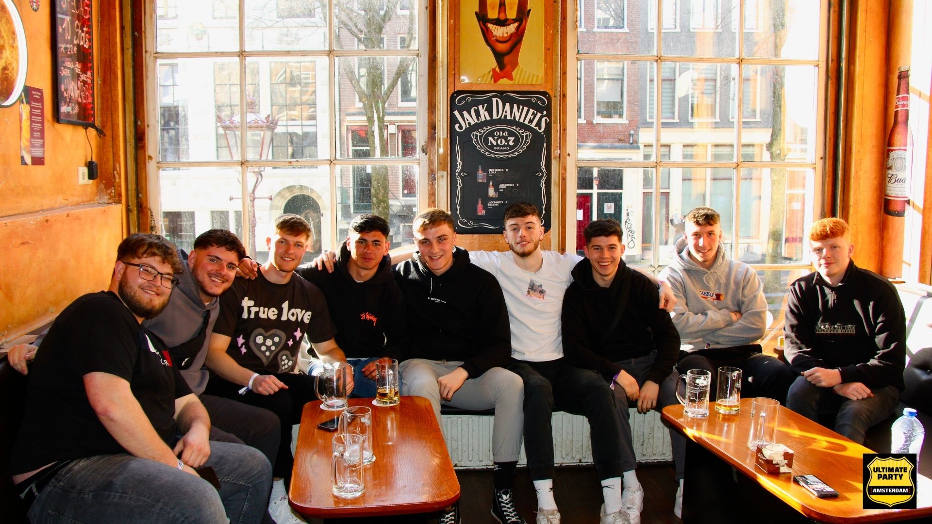 Group of nine young men sitting closely on a bench in a cozy bar with wooden tables and large windows behind them.