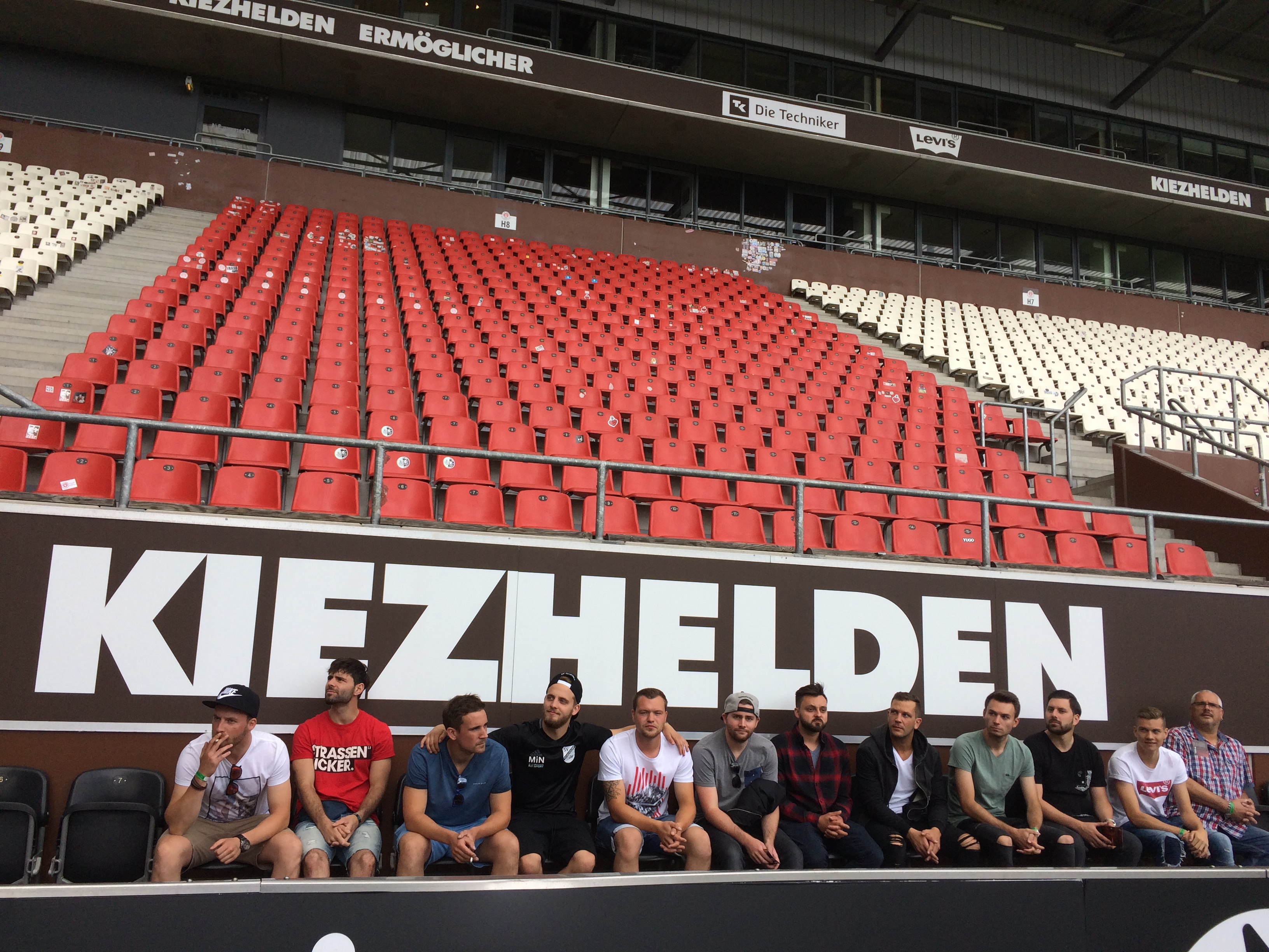 Group of eleven men sitting in front of red and white stadium seats with large 'KIEZHELDEN' sign behind them.
