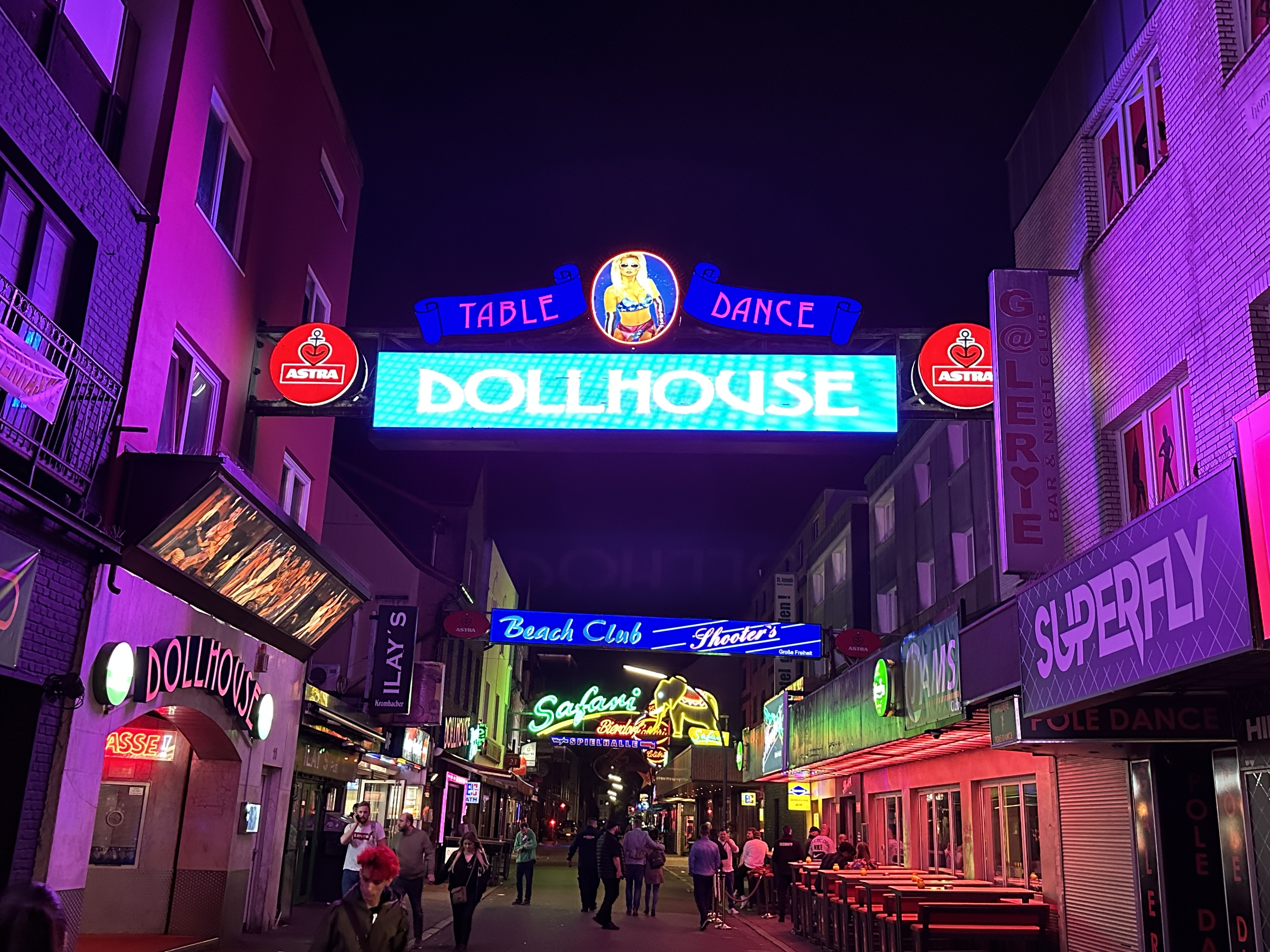 Night street scene with neon signs advertising Dollhouse table dance club and other nightlife venues, with people walking and socializing.