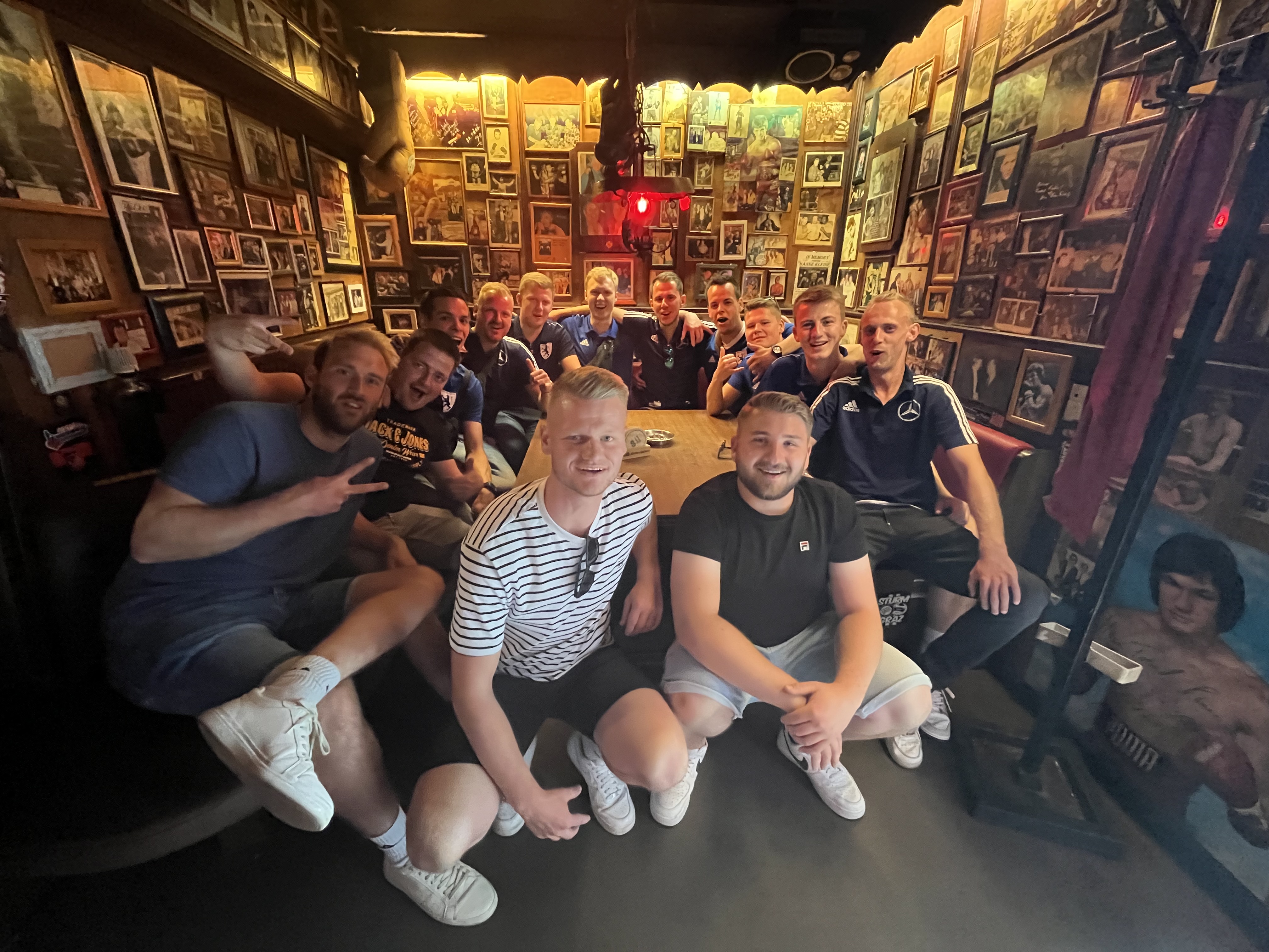 Group of young men posing and smiling inside a dimly lit pub with walls covered in framed pictures.