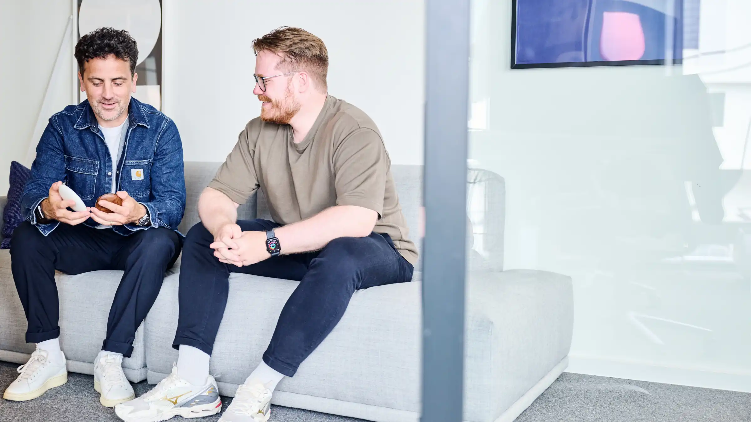 Zwei Männer sitzen auf einem grauen Sofa, einer zeigt dem anderen ein Gerät in der Hand.