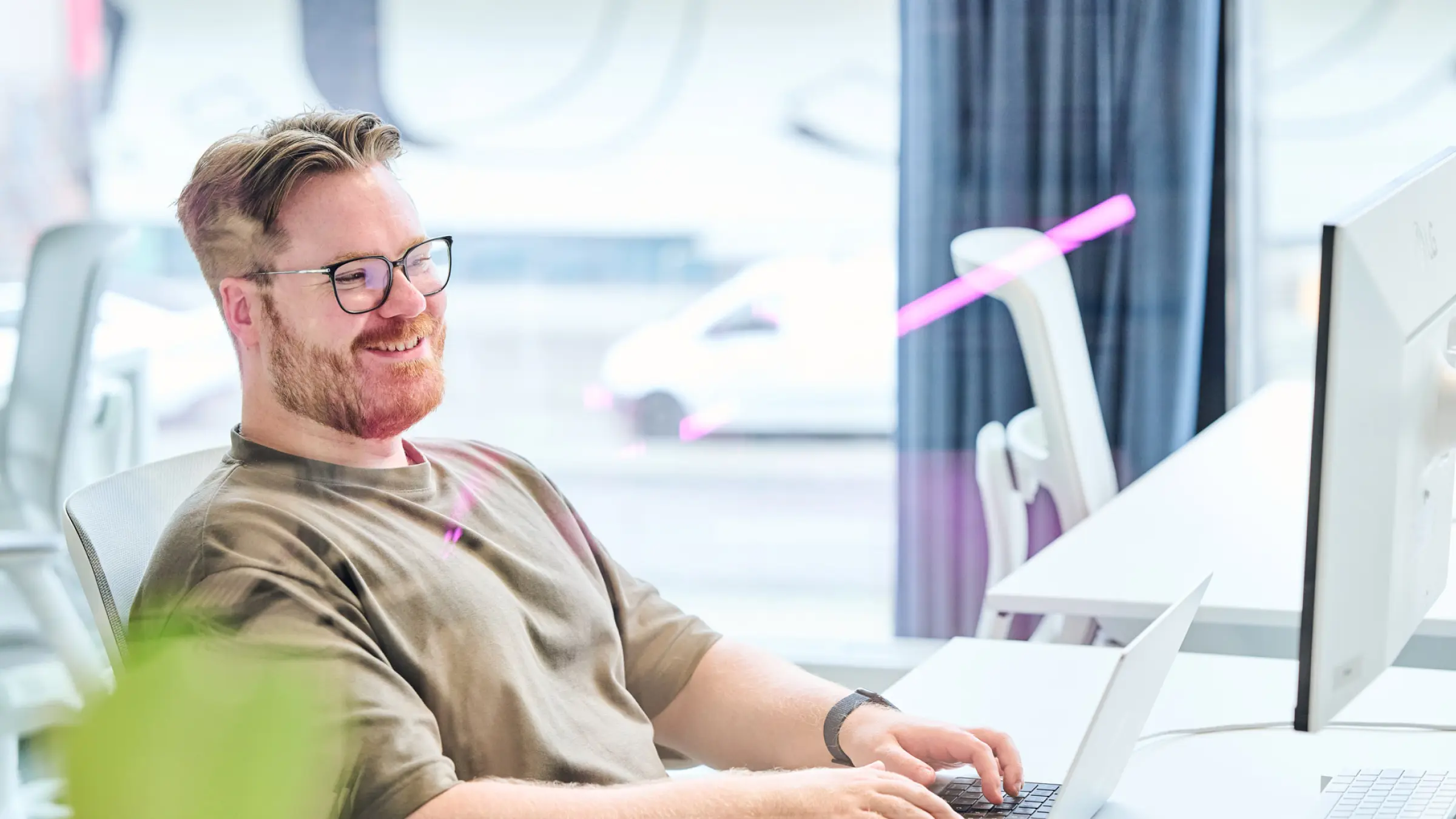 Ein lächelnder Mann mit Brille sitzt an einem Schreibtisch und tippt auf einem Laptop im Büro.