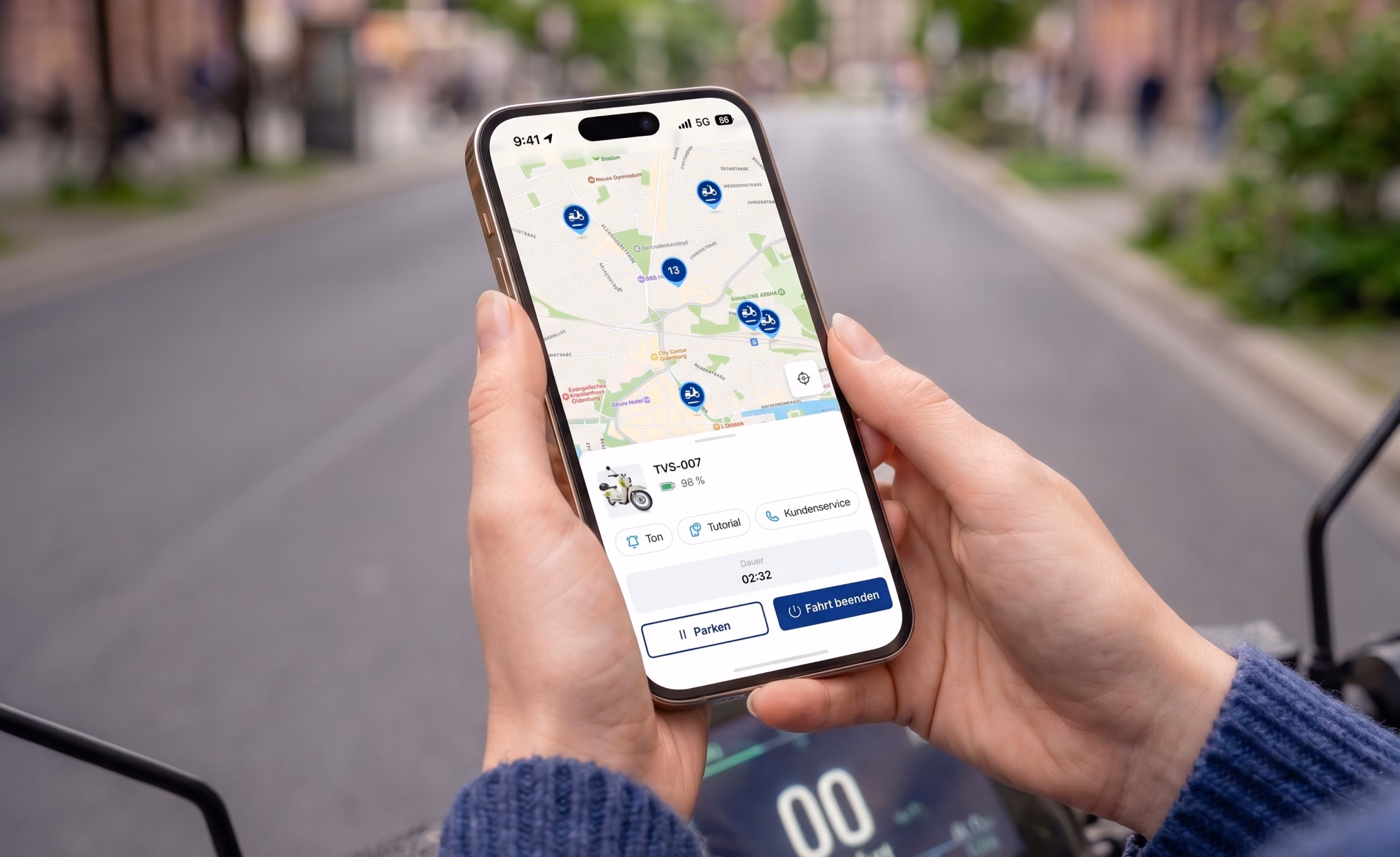 Hände halten ein Smartphone mit der TVS E-Scooter-App: Kartenansicht mit Fahrzeug-Markern und laufender Fahrt