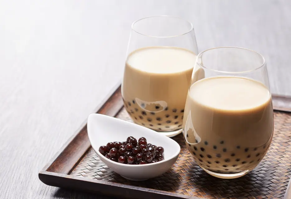 Two glasses of traditional milk tea served with a side dish of black tapioca pearls on a wooden tray