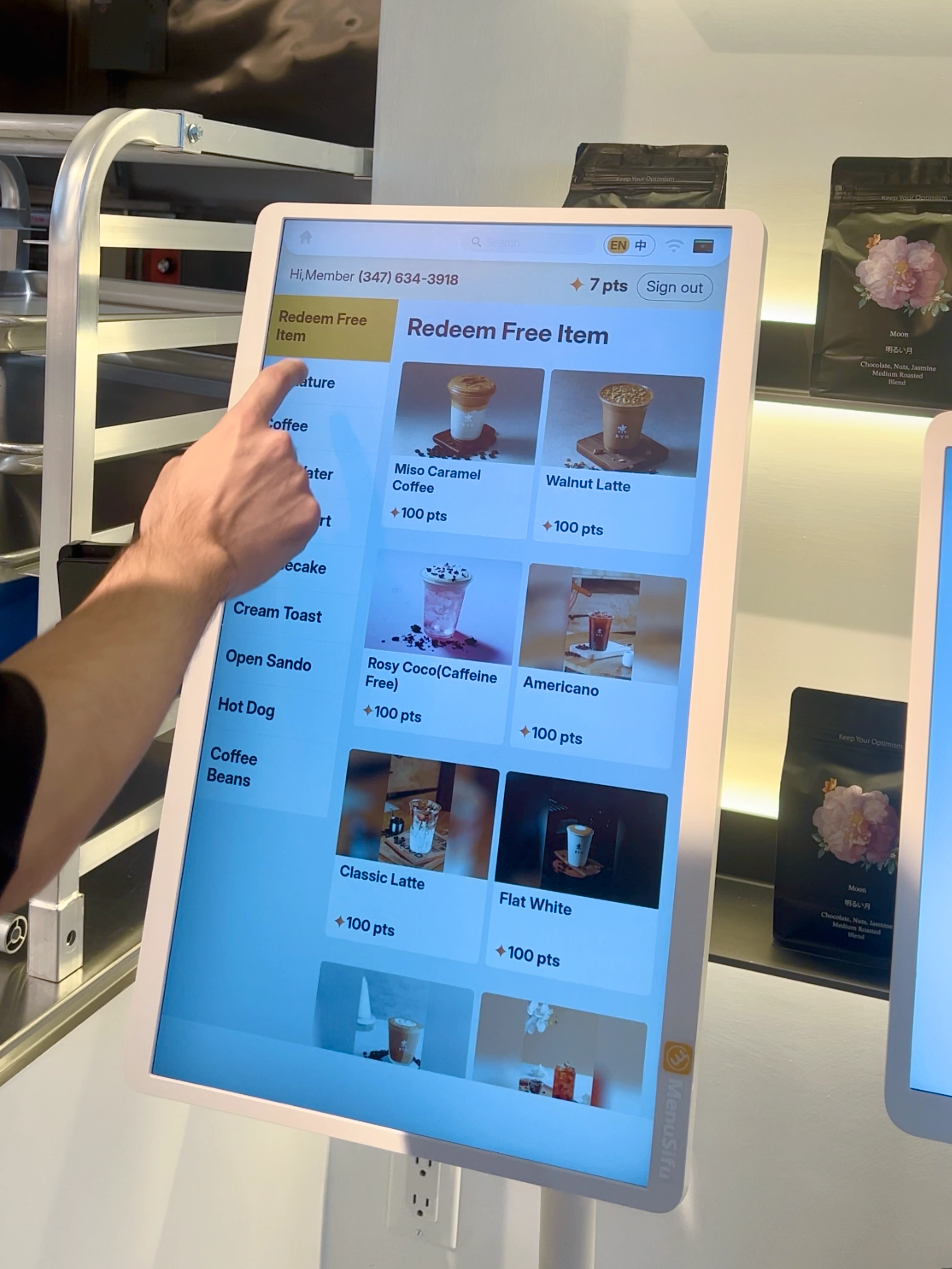 顾客手指正在操作MenuSifu自助点餐机屏幕,选择使用积分兑换饮品,界面显示多种咖啡和饮品选项。