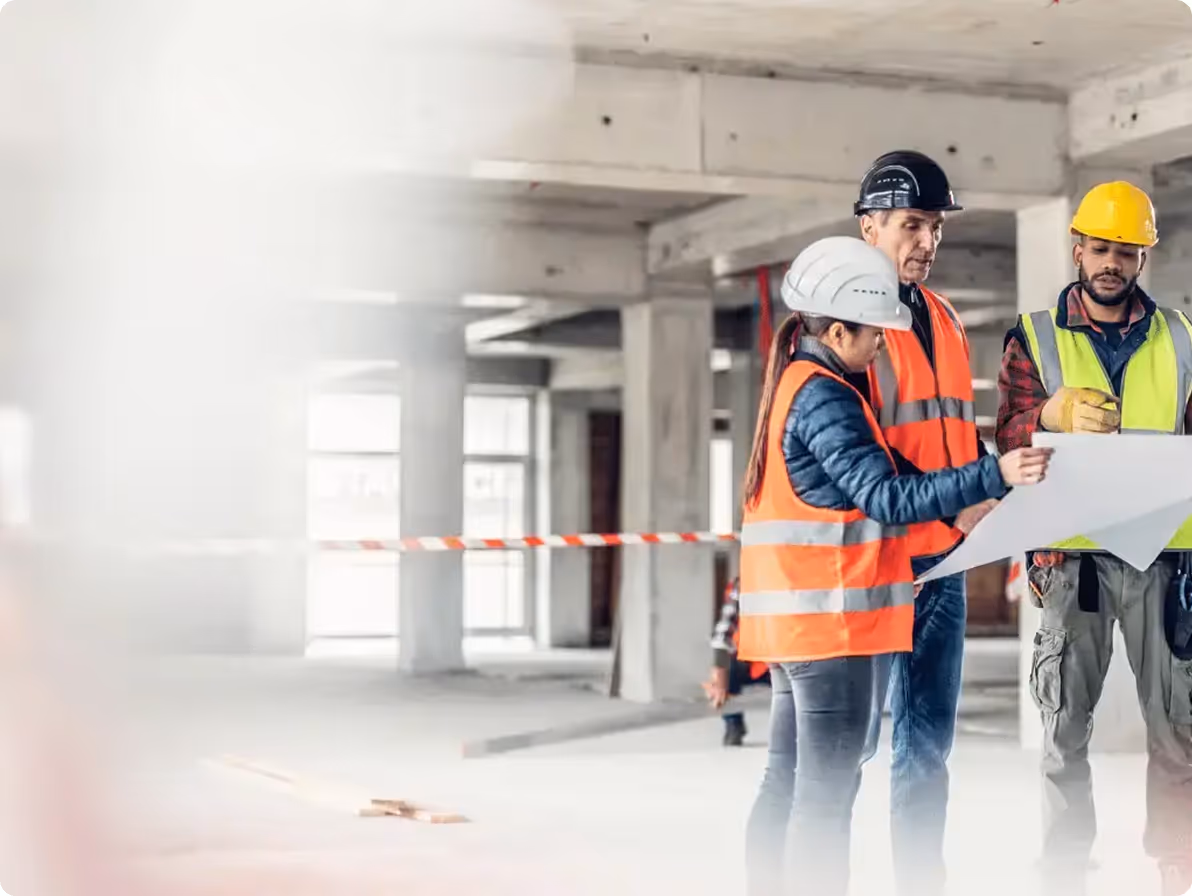 Three construction workers in safety helmets and vests reviewing a blueprint inside an unfinished building.