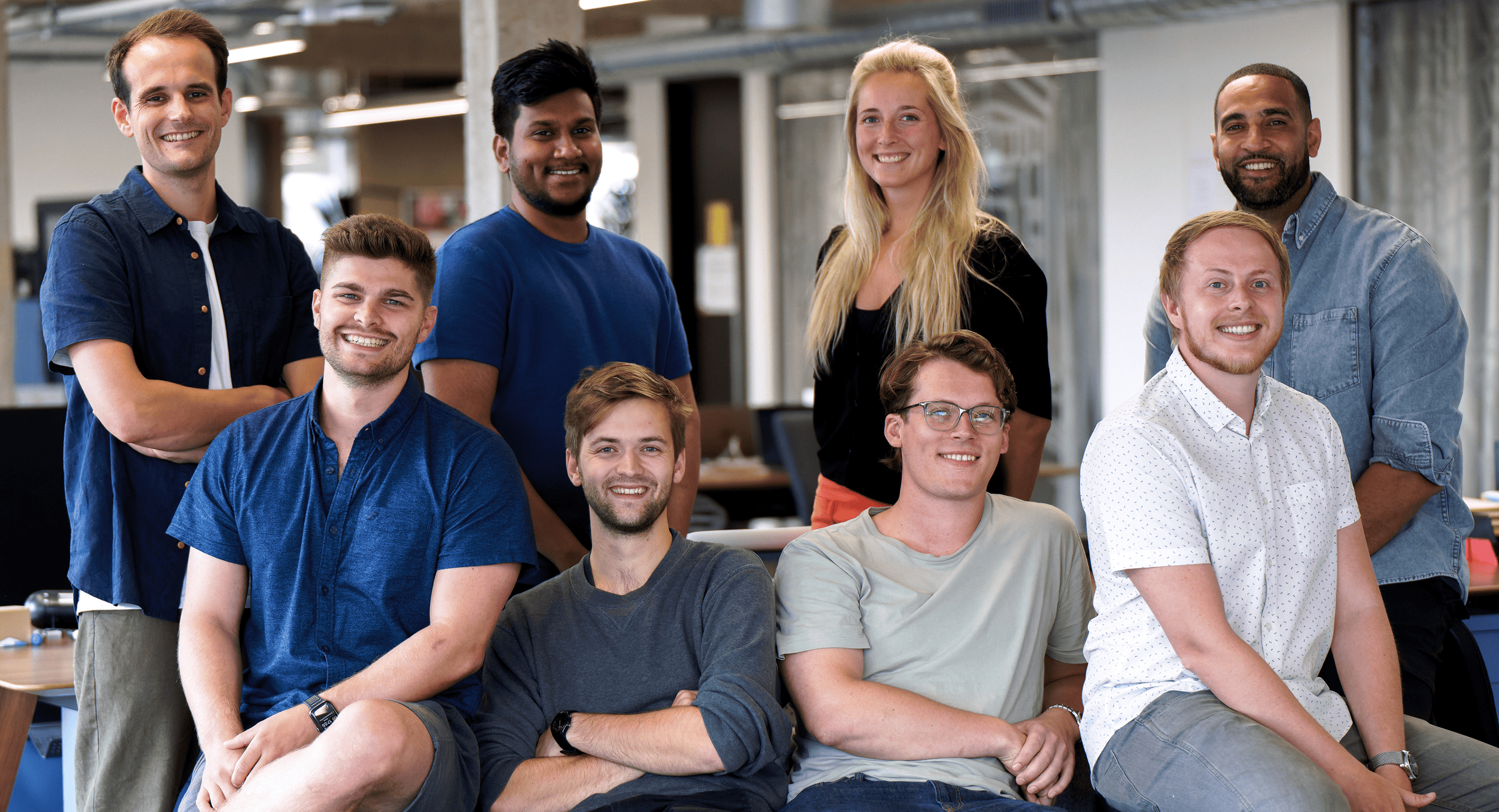 Eight young adults smiling at the camera in a modern office setting, with five seated in front and three standing behind.