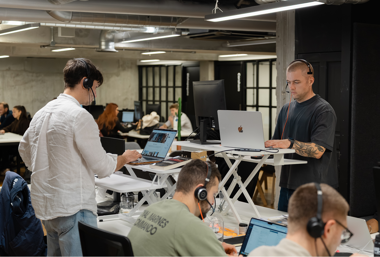 Four men wearing headsets working on laptops in a modern office with standing desks and industrial-style ceiling.