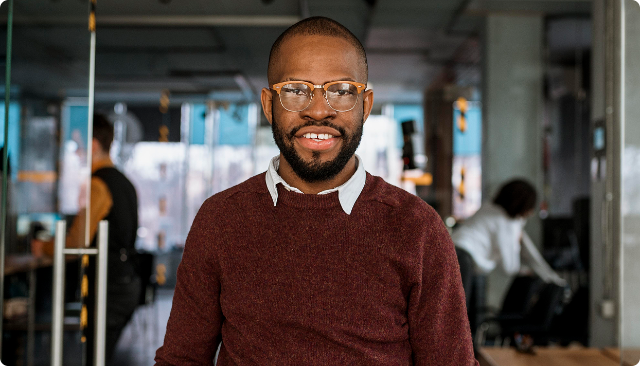 Smiling man wearing glasses and a maroon sweater in a modern office setting.