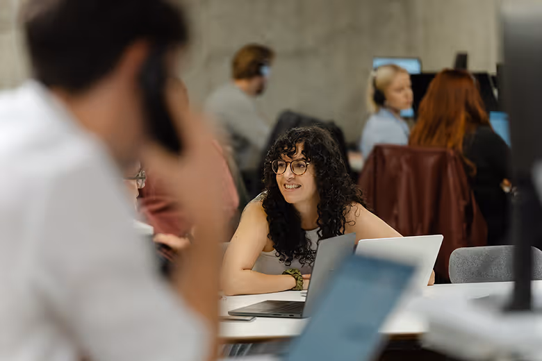 A group of five coworkers in an office celebrating a birthday with a chocolate cake with lit candles and a gift box.