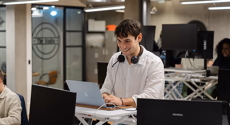 A group of five coworkers in an office celebrating a birthday with a chocolate cake with lit candles and a gift box.