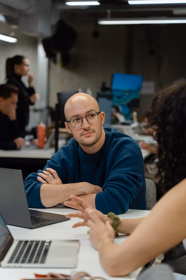 A group of five coworkers in an office celebrating a birthday with a chocolate cake with lit candles and a gift box.