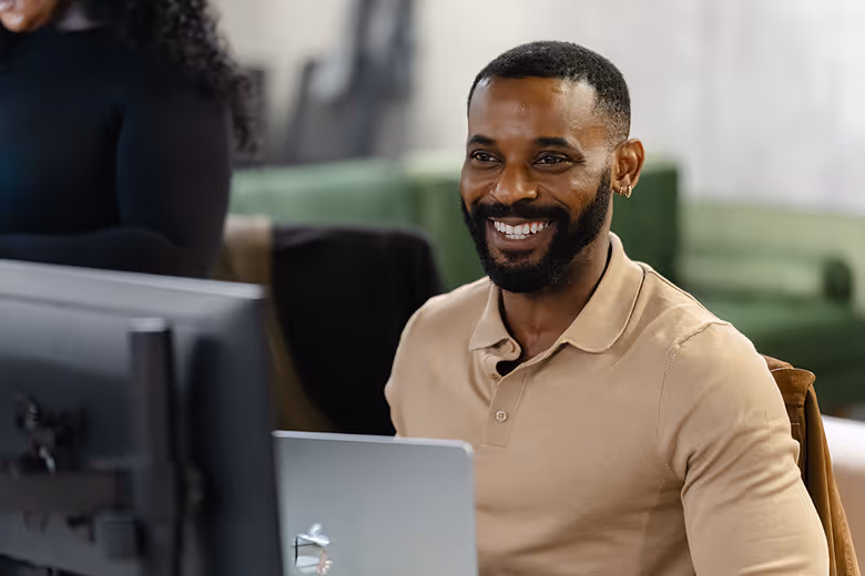 A group of five coworkers in an office celebrating a birthday with a chocolate cake with lit candles and a gift box.
