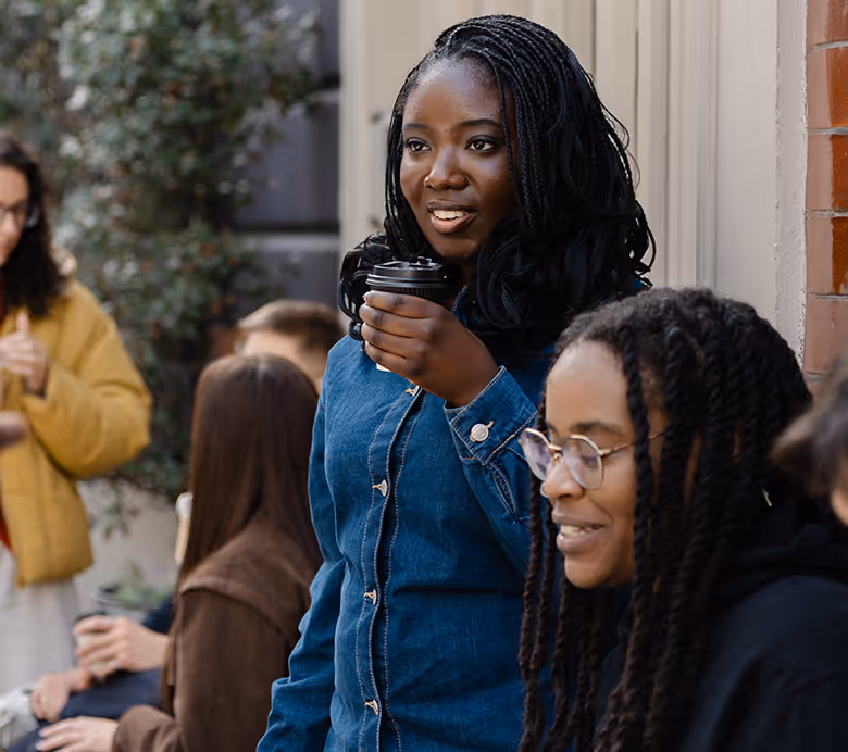 A group of five coworkers in an office celebrating a birthday with a chocolate cake with lit candles and a gift box.