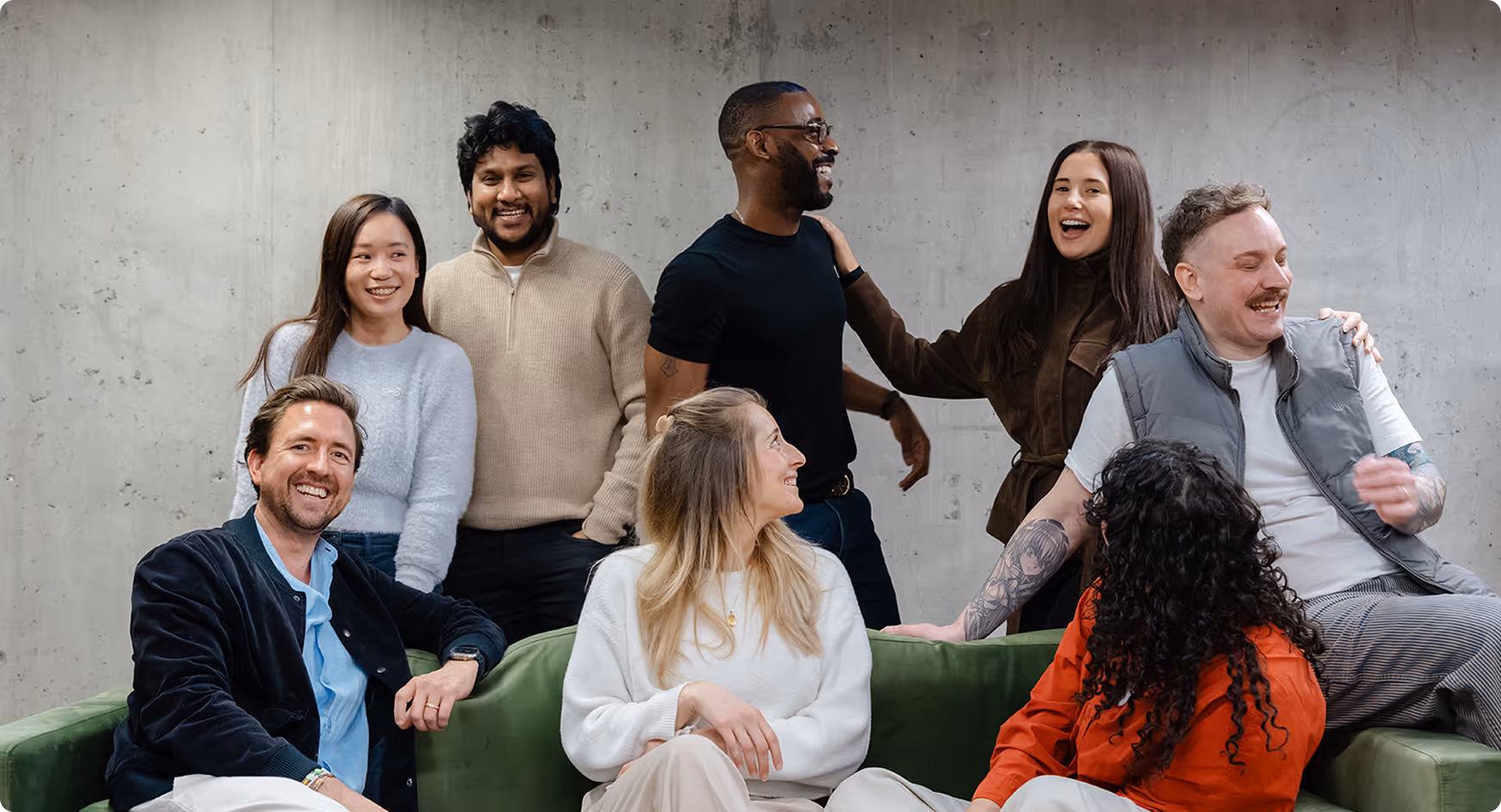 Diverse group of eight smiling people casually interacting in front of a grey concrete wall with some sitting on a green sofa.
