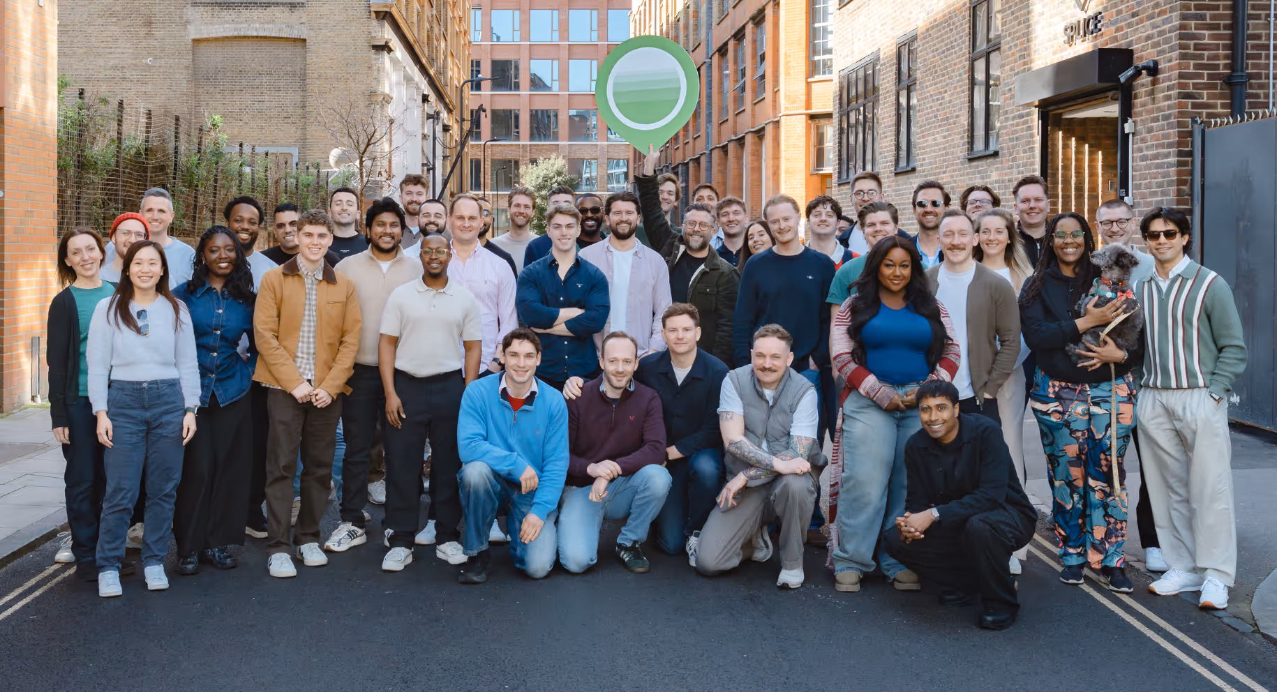 Large diverse group of people posing for a photo in an urban street setting, one person holds a green and white circular sign above their head.