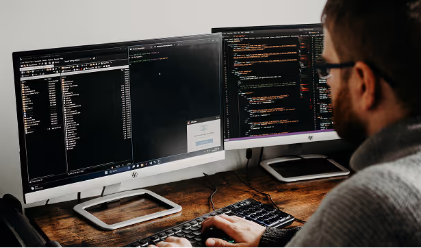 Software engineer working at a desk with dual monitors displaying code and development tools.