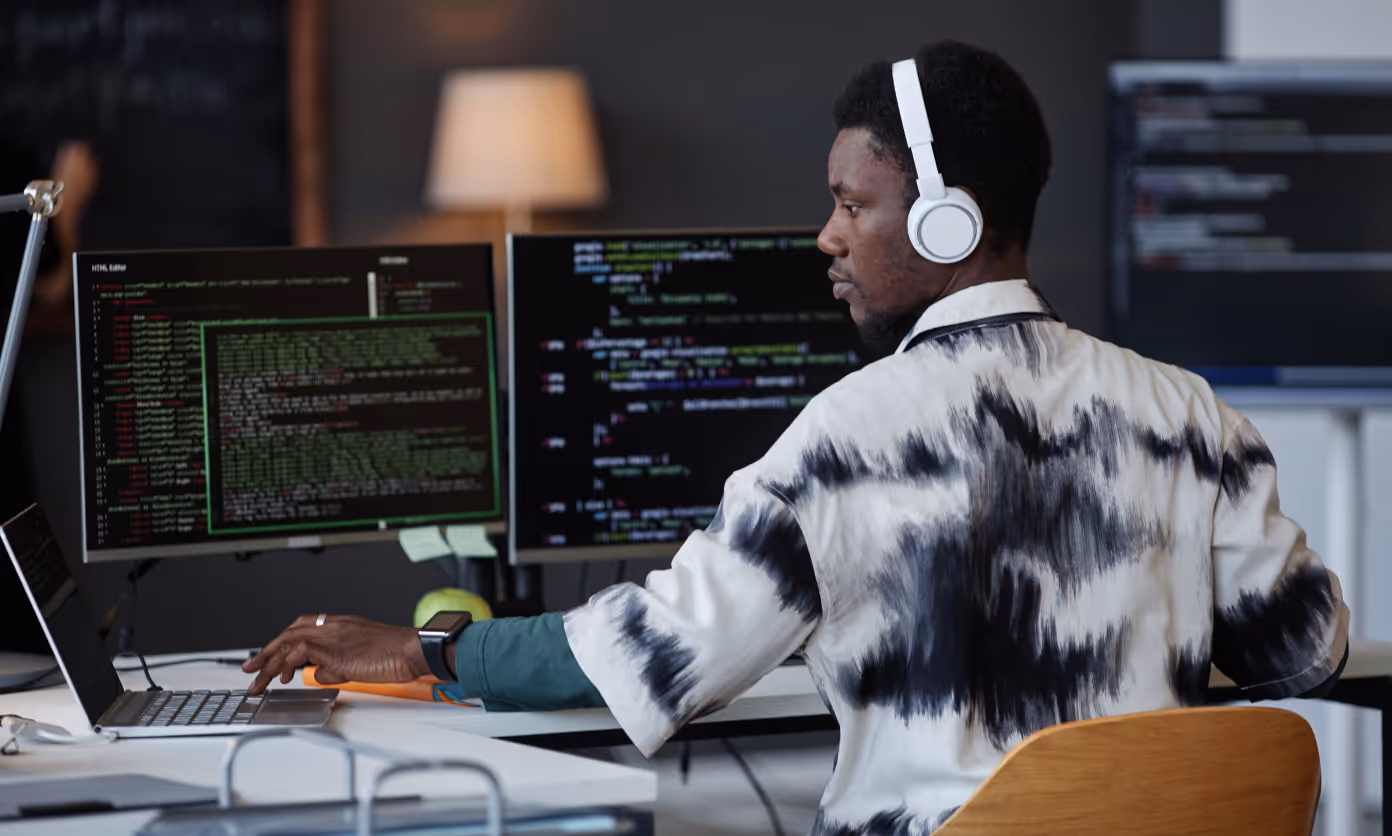 Man wearing white headphones and a tie-dye shirt working on a laptop and desktop monitors displaying code in a modern office.