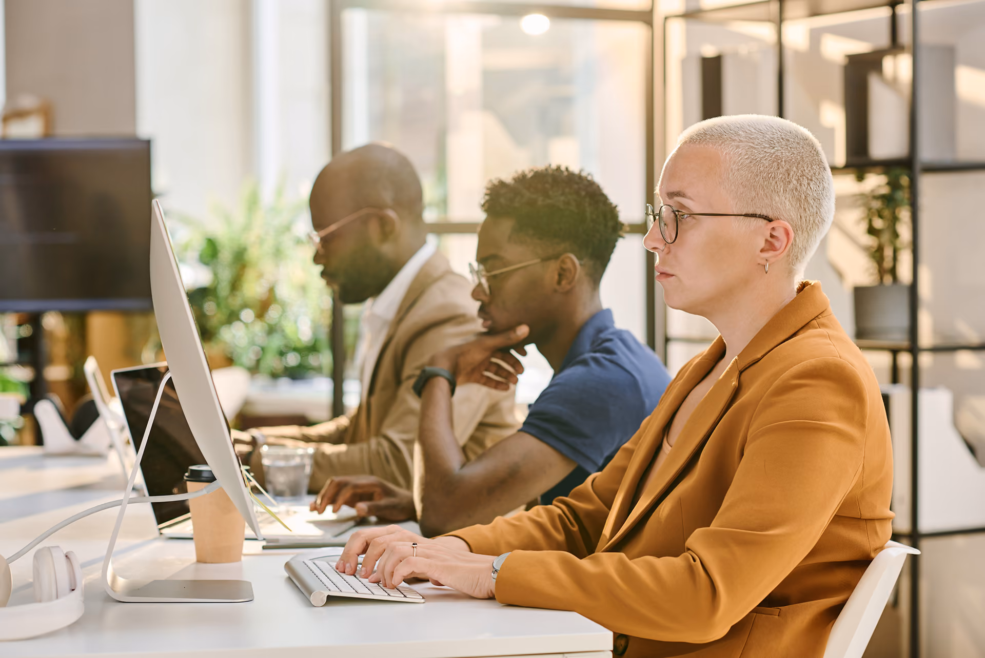 Three diverse professionals focused, working side-by-side on computers at a bright modern office desk.