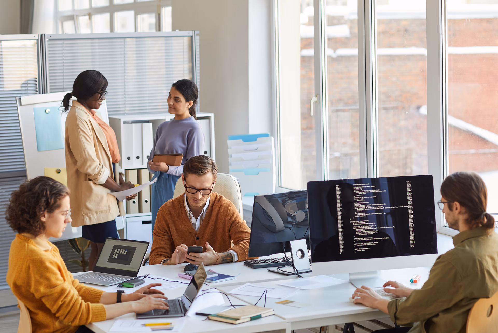 Five diverse coworkers collaborate and work on laptops and a desktop with code in a bright office.