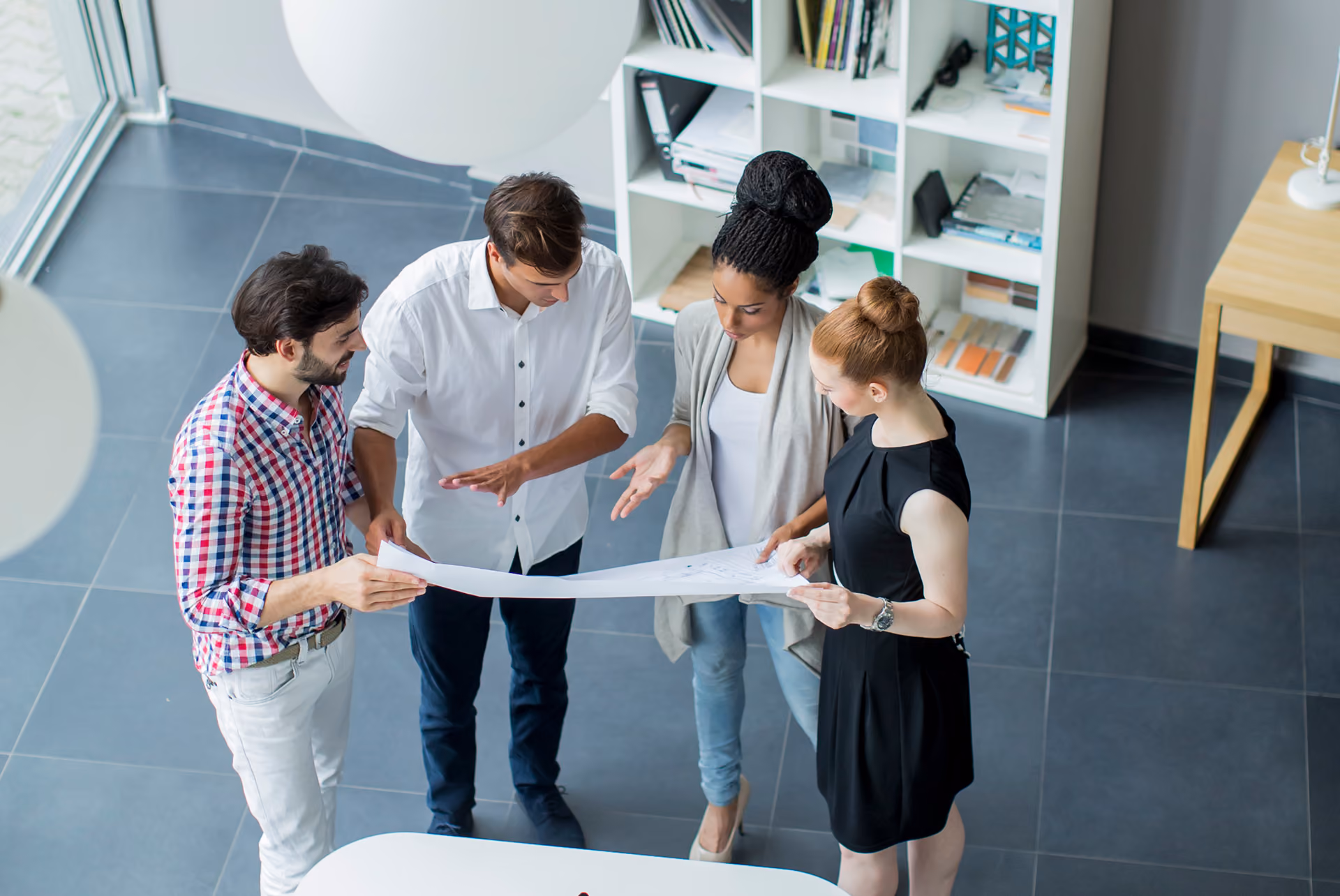 Four diverse professionals standing in an office discussing a large sheet of paper.