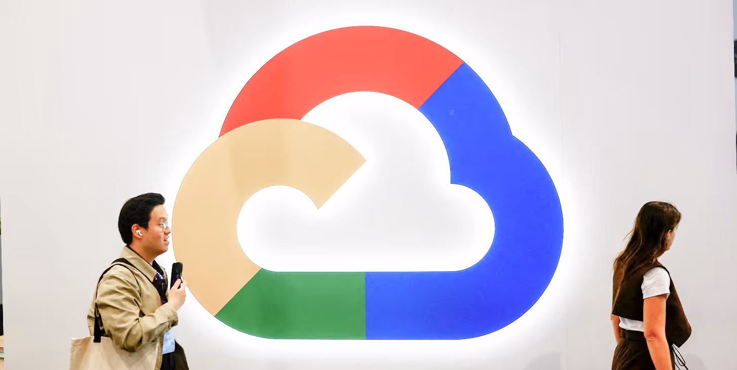 Two people walking past a large colorful cloud-shaped logo from Google on a white wall.