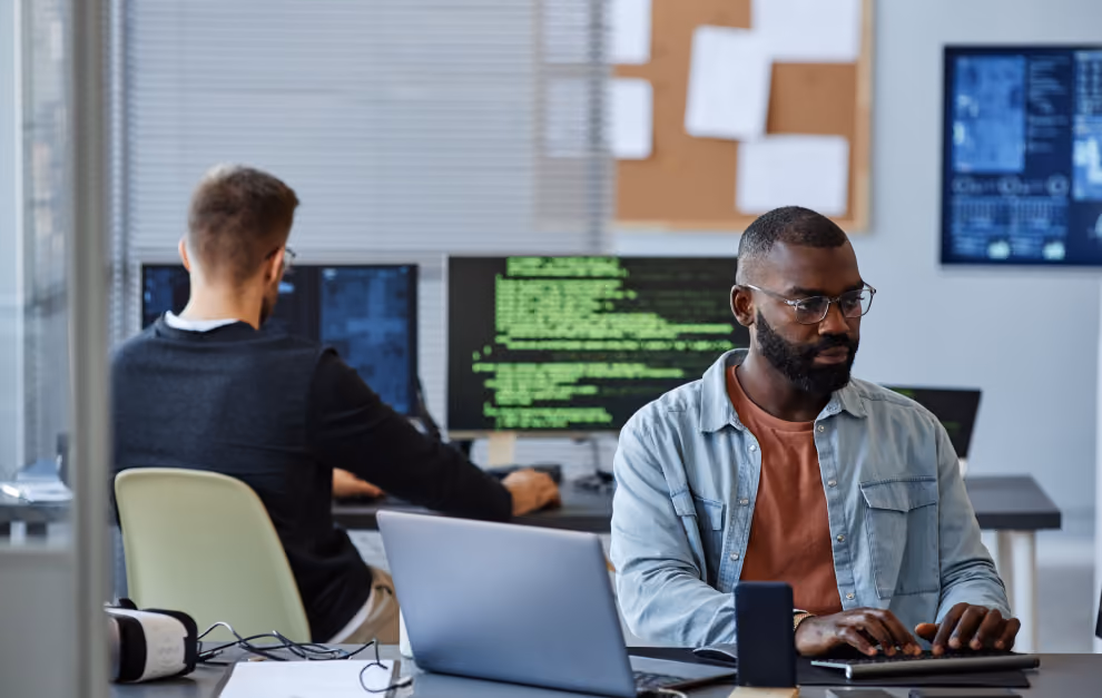 Two men working at desks in a modern office; the man in the foreground wears glasses and a light denim shirt while typing on a laptop, the man in the background is focused on dual monitors displaying code and data.