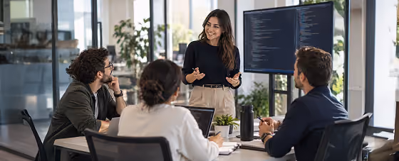 A woman standing and presenting code on a large screen to three seated coworkers around a conference table in a modern office.