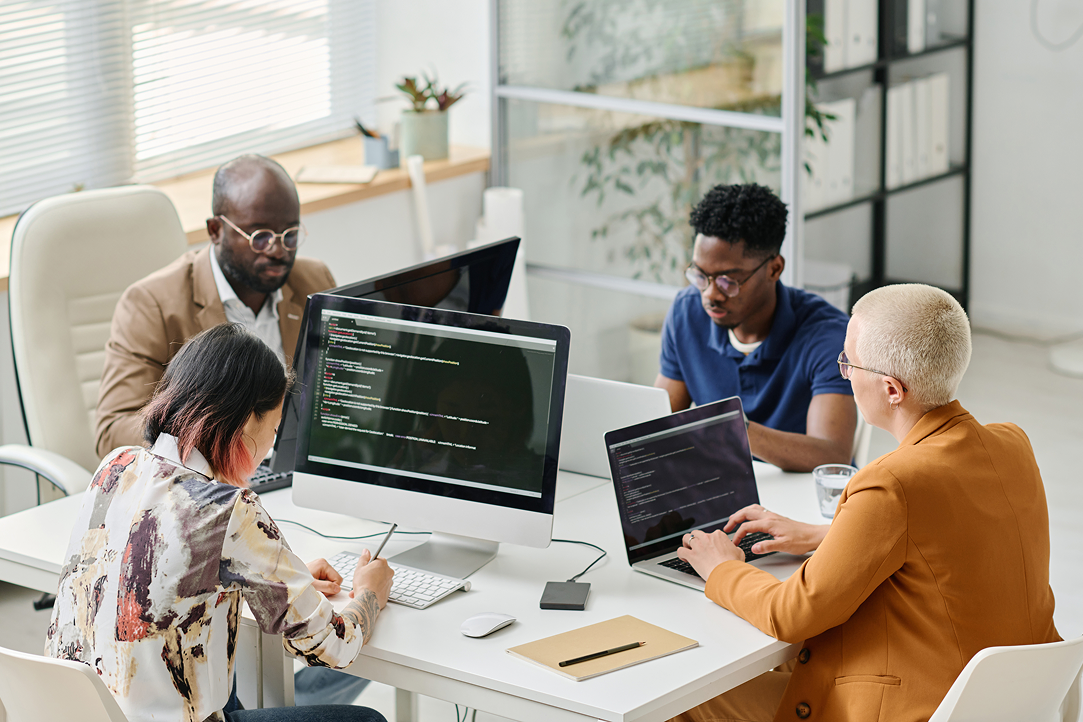 Diverse software development team collaborating at a modern office table, writing code on laptops and desktop computers while working on AI and data-driven solutions for private equity firms.