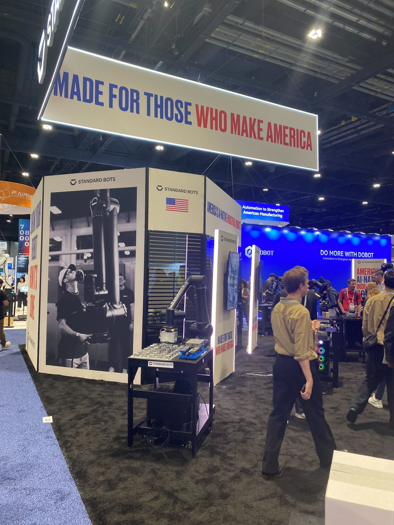 Standard Bots booth at Automate 2026 featuring a robotic arm demonstration, overhead "Made for Those Who Make America" signage, and attendees on the exhibition floor.