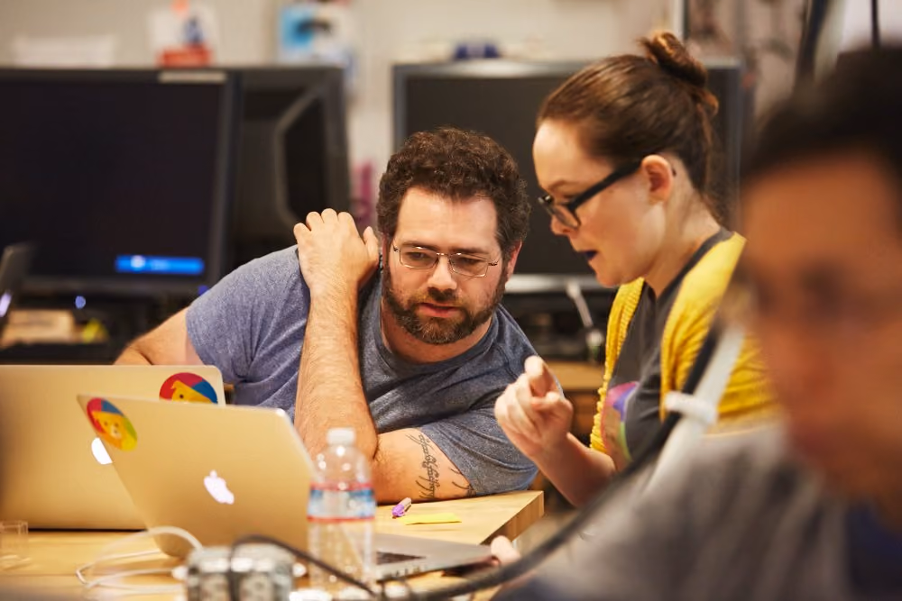 Two colleagues collaborating at a desk, discussing code on their laptops in a modern office workspace. Source: Google